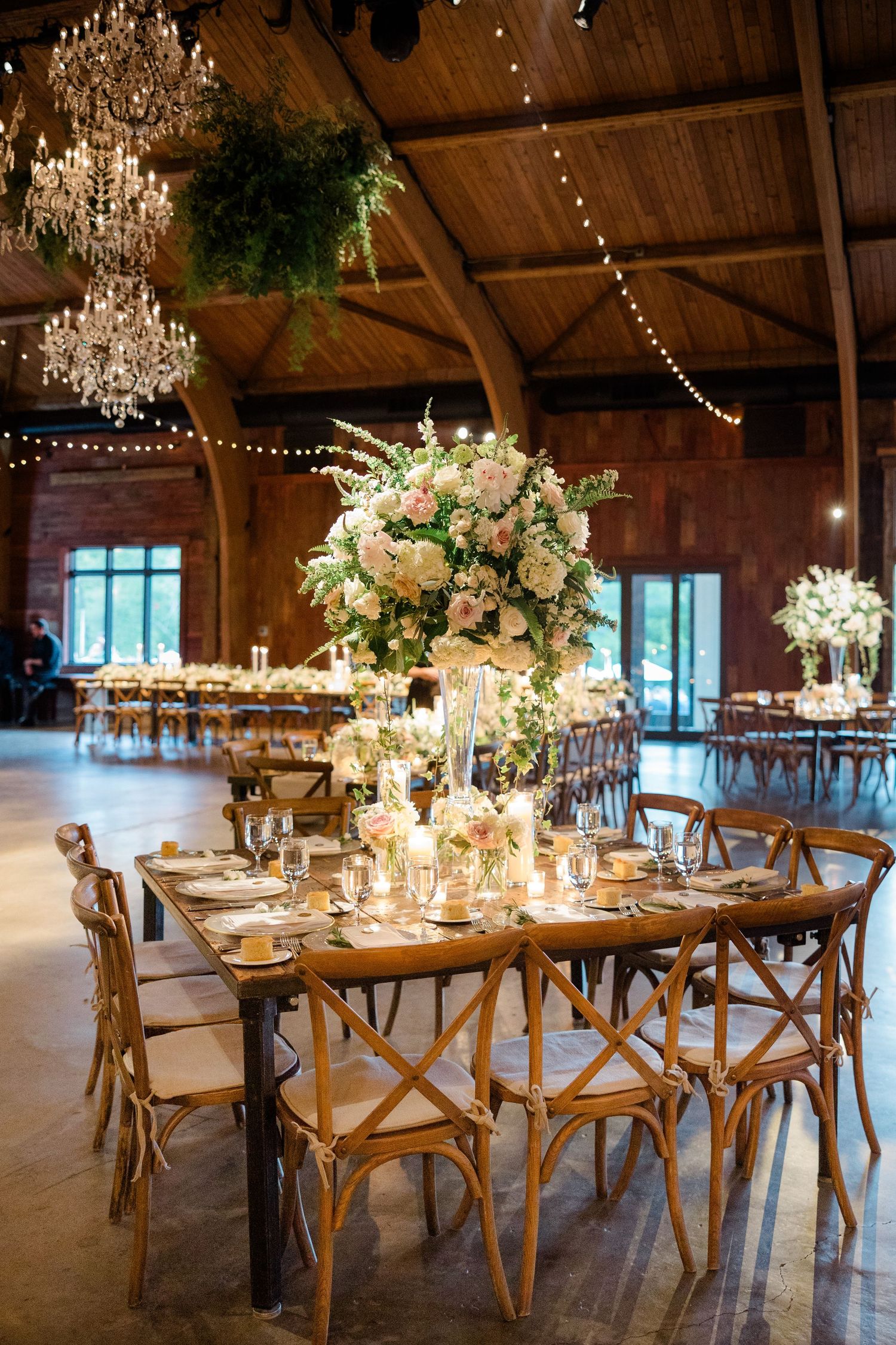 Elegant wedding reception table with towering floral centerpiece, crystal chandeliers, and rustic cross-back chairs.