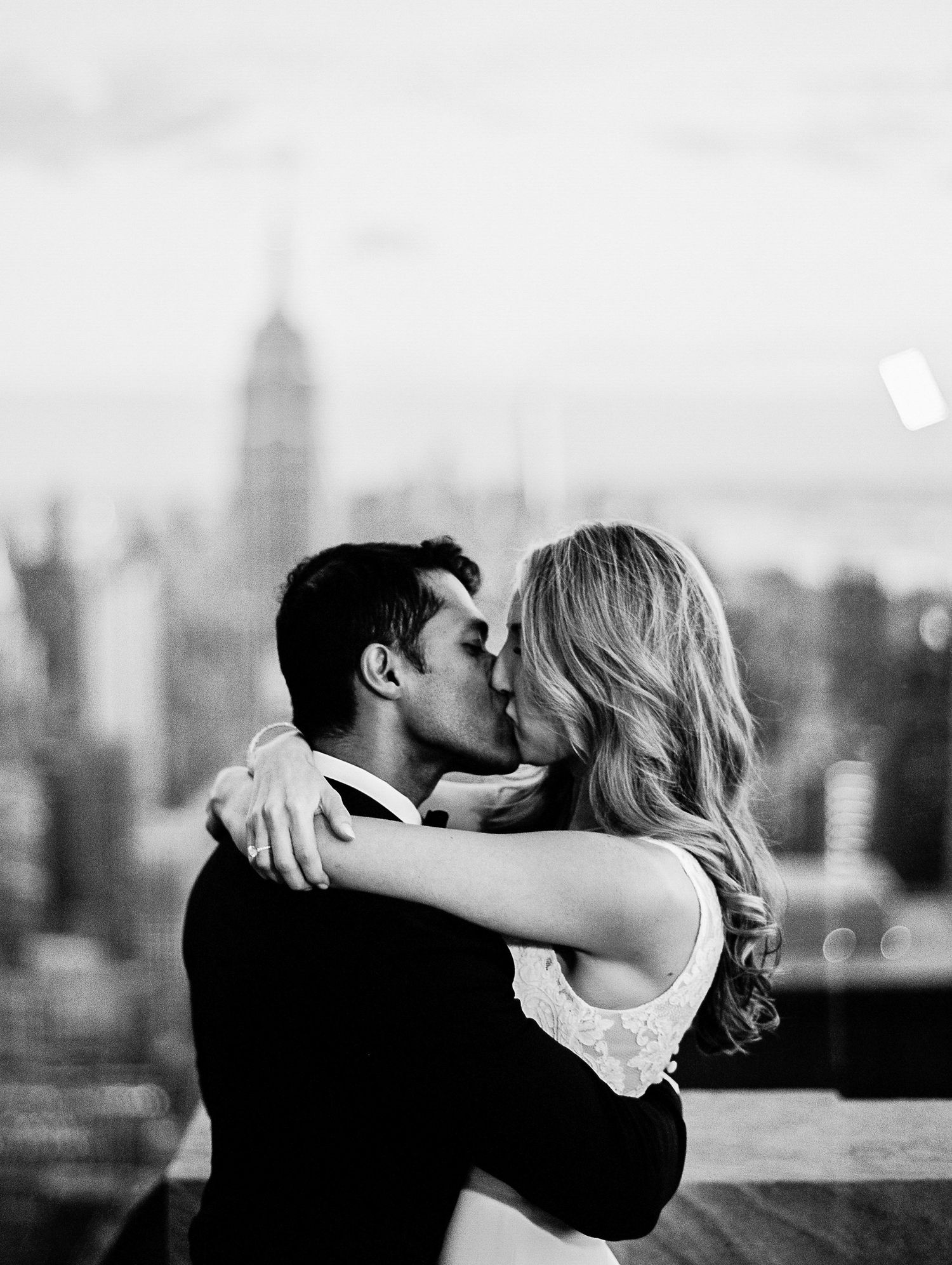 A romantic black and white cityscape photo shows a couple embracing and kissing with the NYC skyline in the background.