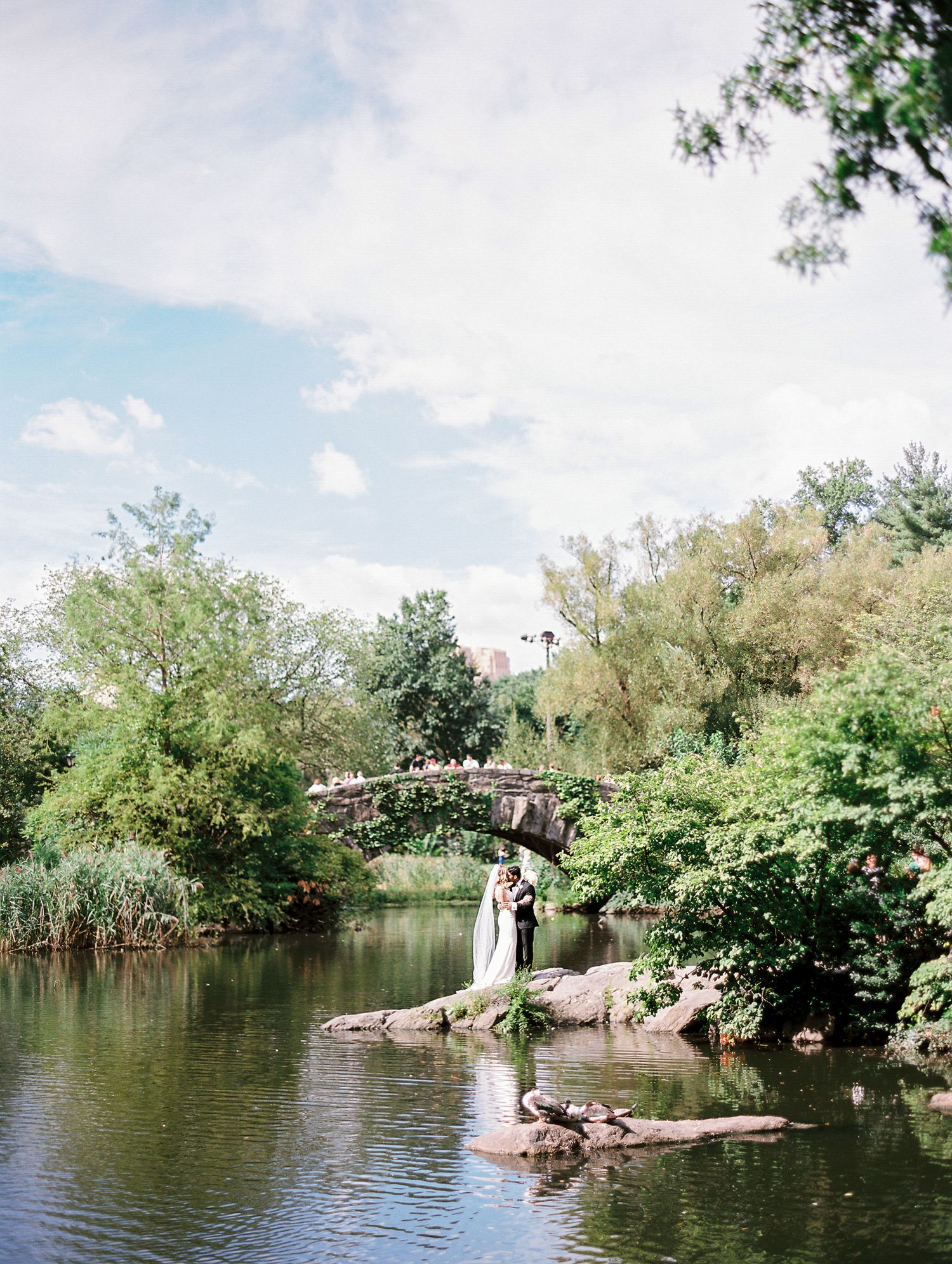 A scenic pond with an arched stone bridge surrounded by lush trees and greenery on a sunny summer day.