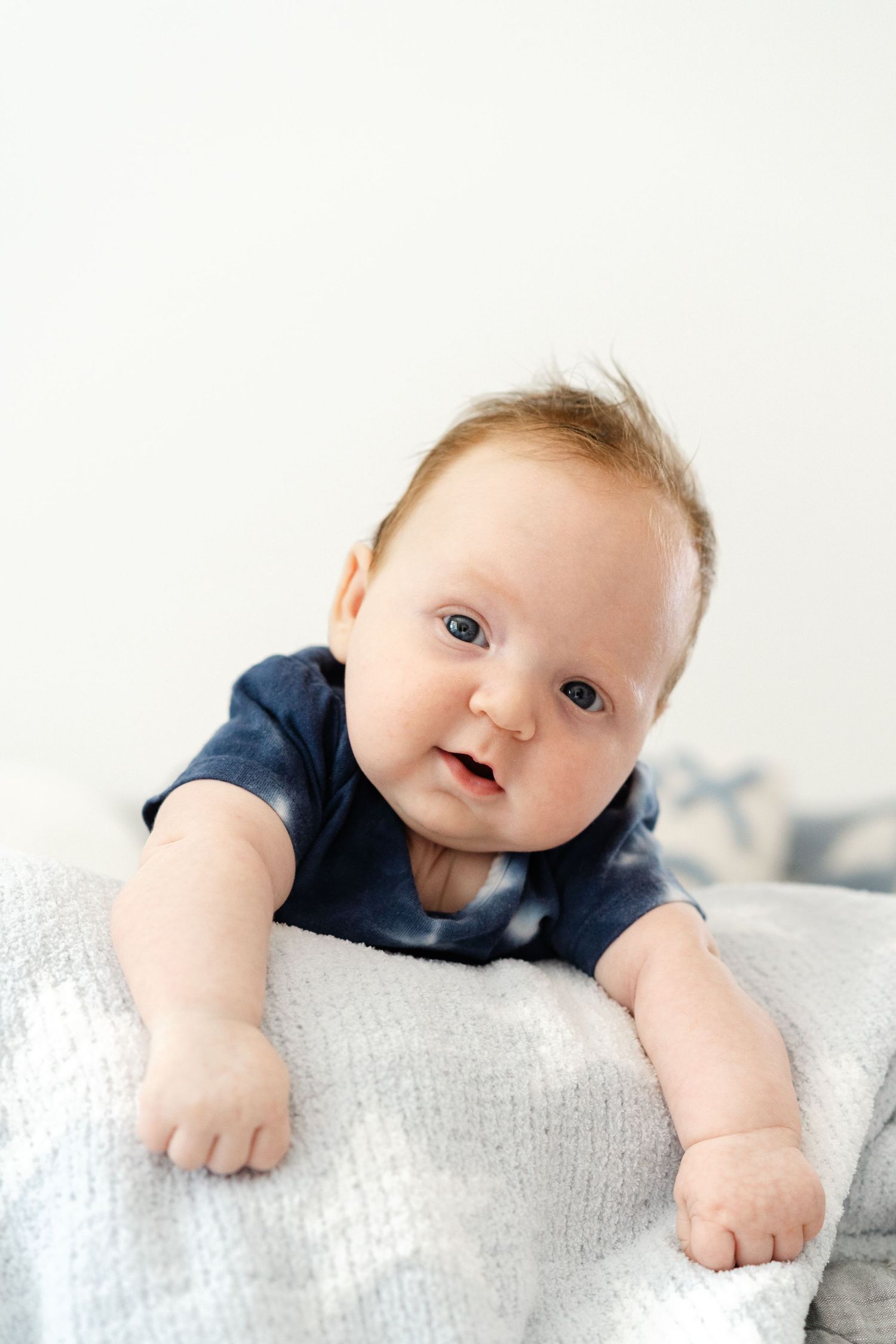 A baby in a navy blue shirt lies on a light gray cushion while looking at the camera.