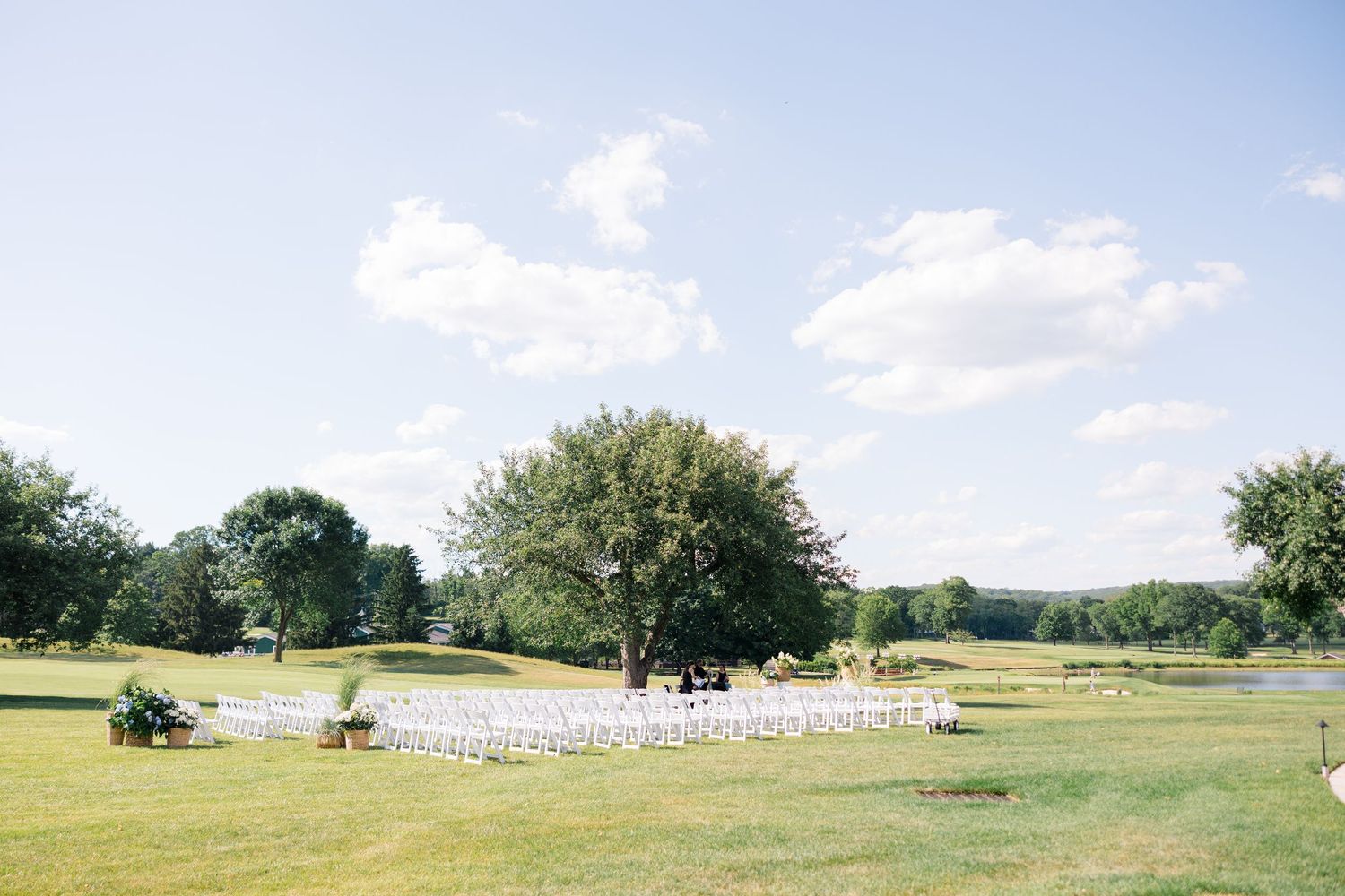 Outdoor wedding ceremony setup with white chairs arranged under a large tree on a lush green golf course lawn.