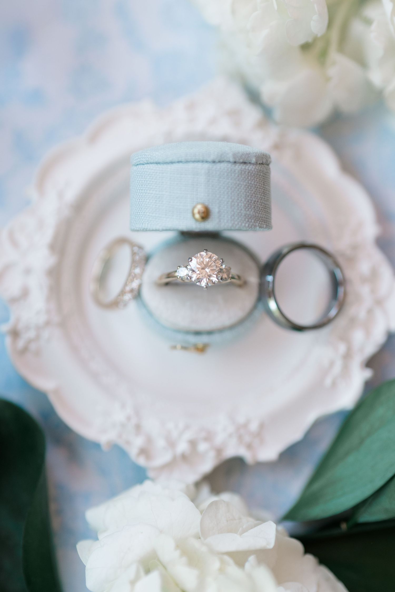 Elegant wedding rings displayed in a blue velvet ring box on a vintage pink decorative plate with white flowers.