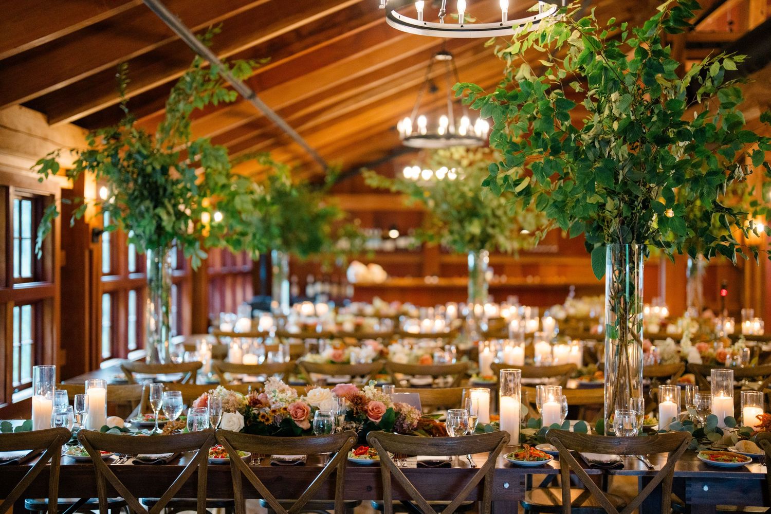 Elegant wedding reception hall with tall greenery centerpieces, long tables, candles, and wooden ceiling in a rustic venue.