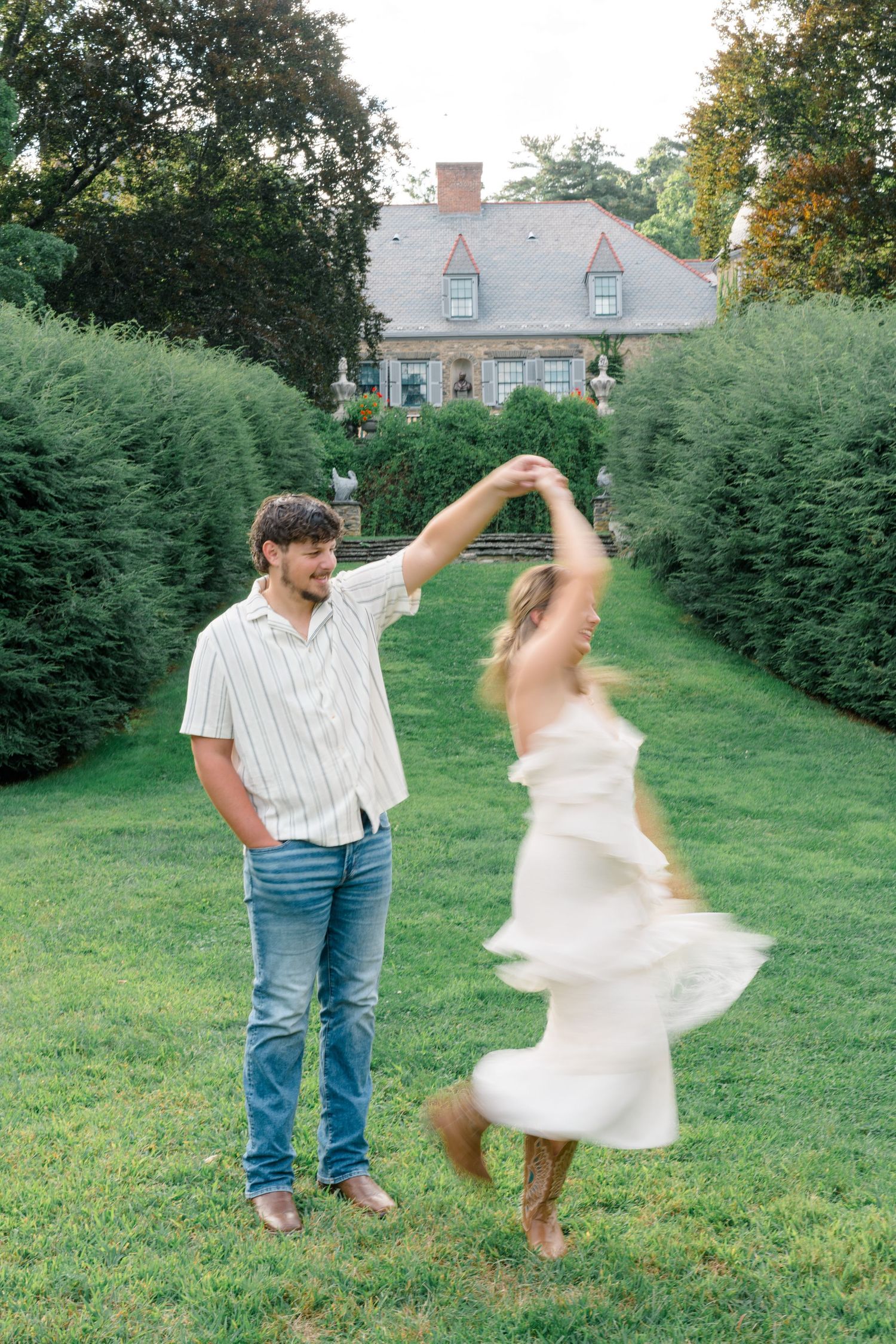 A man in jeans twirls a woman in a white dress on a green lawn beside tall hedges with an elegant manor house behind them.