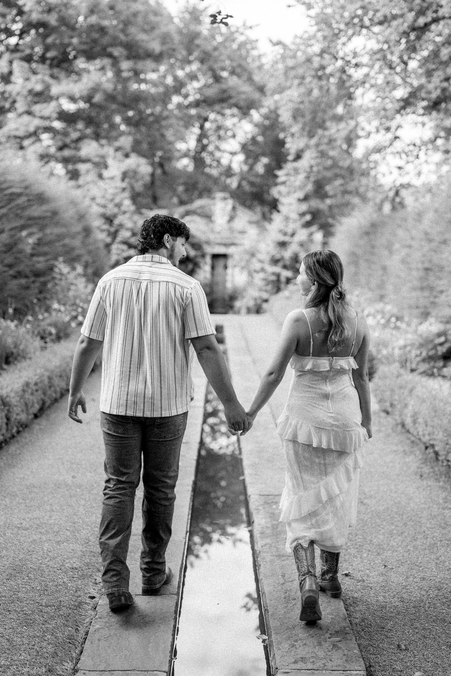 Black and white photo of couple holding hands walking down garden path toward stone structure.