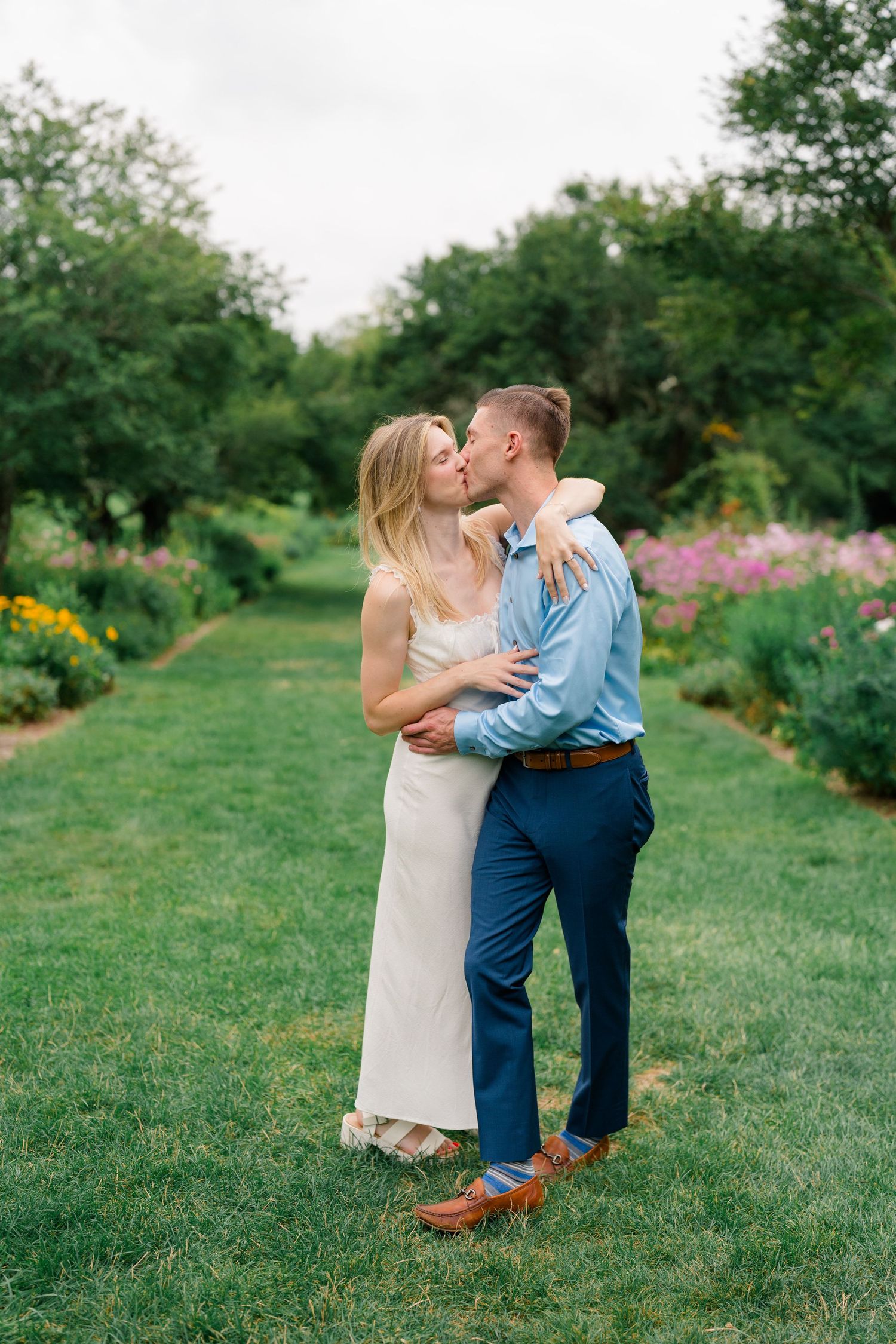 Couple sharing a romantic kiss in a lush garden, woman in white dress, man in blue suit.