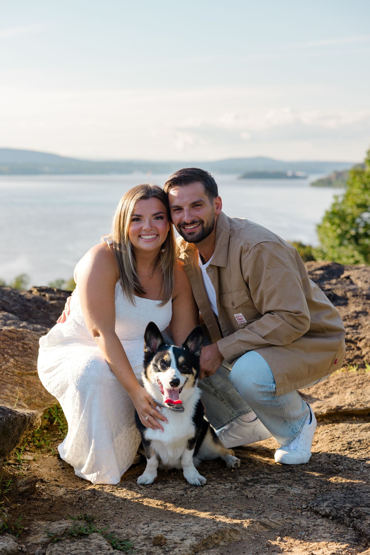 Smiling couple crouching with their black and white Corgi dog on rocky overlook with lake and hills in background.