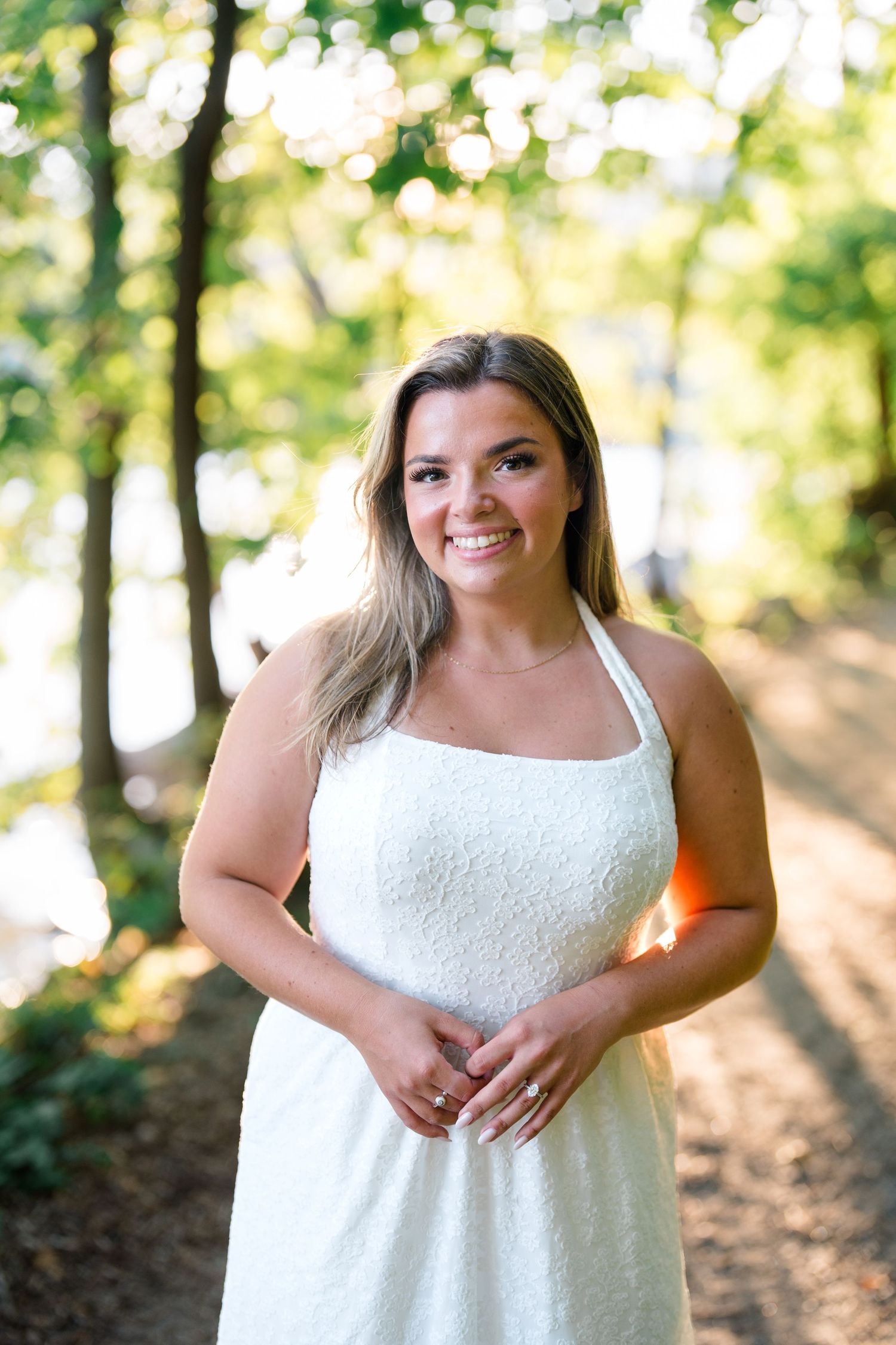 Smiling bride in white lace halter dress poses outdoors among sunlit trees on a forest path.