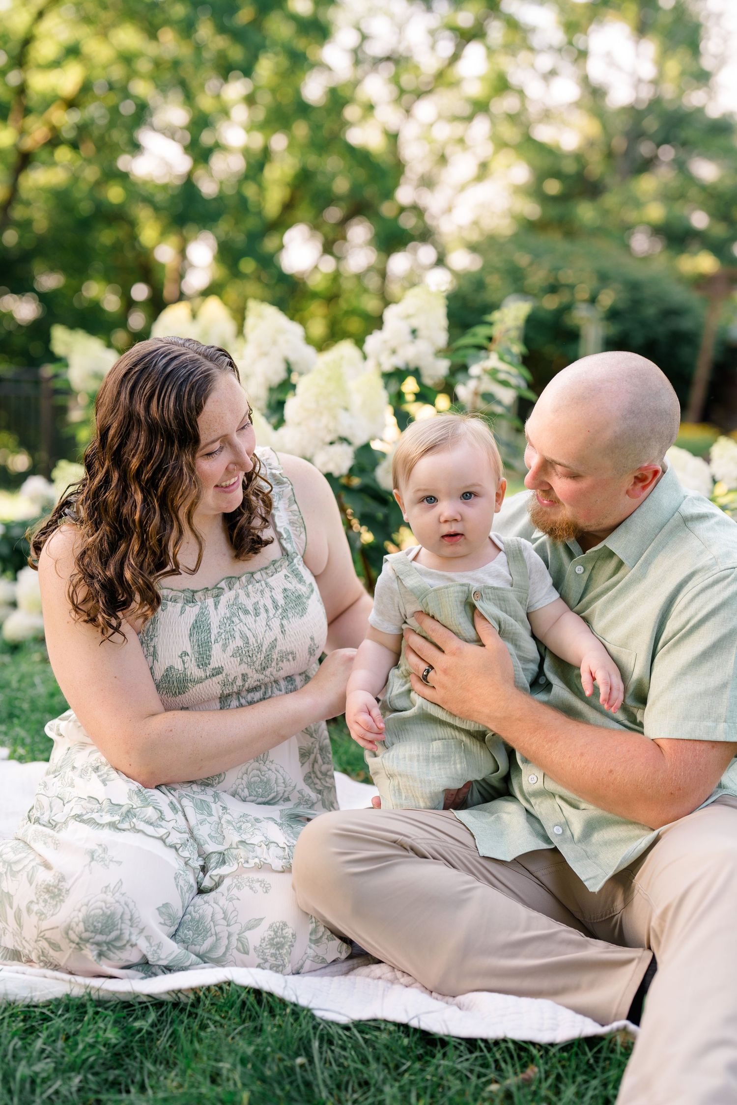 Family of three sitting on blanket in garden with white hydrangeas, parents adoring baby boy.