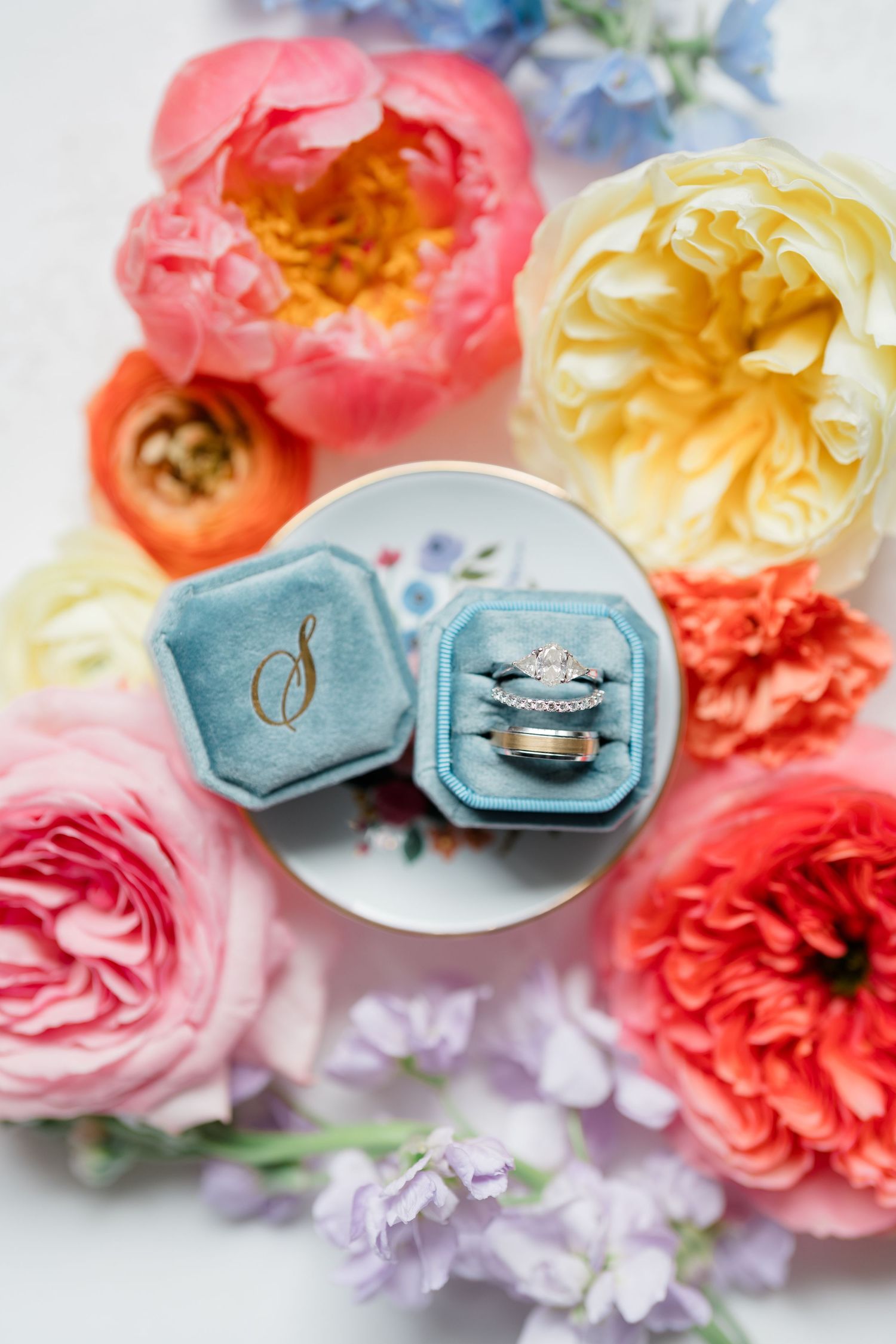 Diamond wedding rings nestled in a blue velvet ring box surrounded by coral, pink and yellow garden roses.
