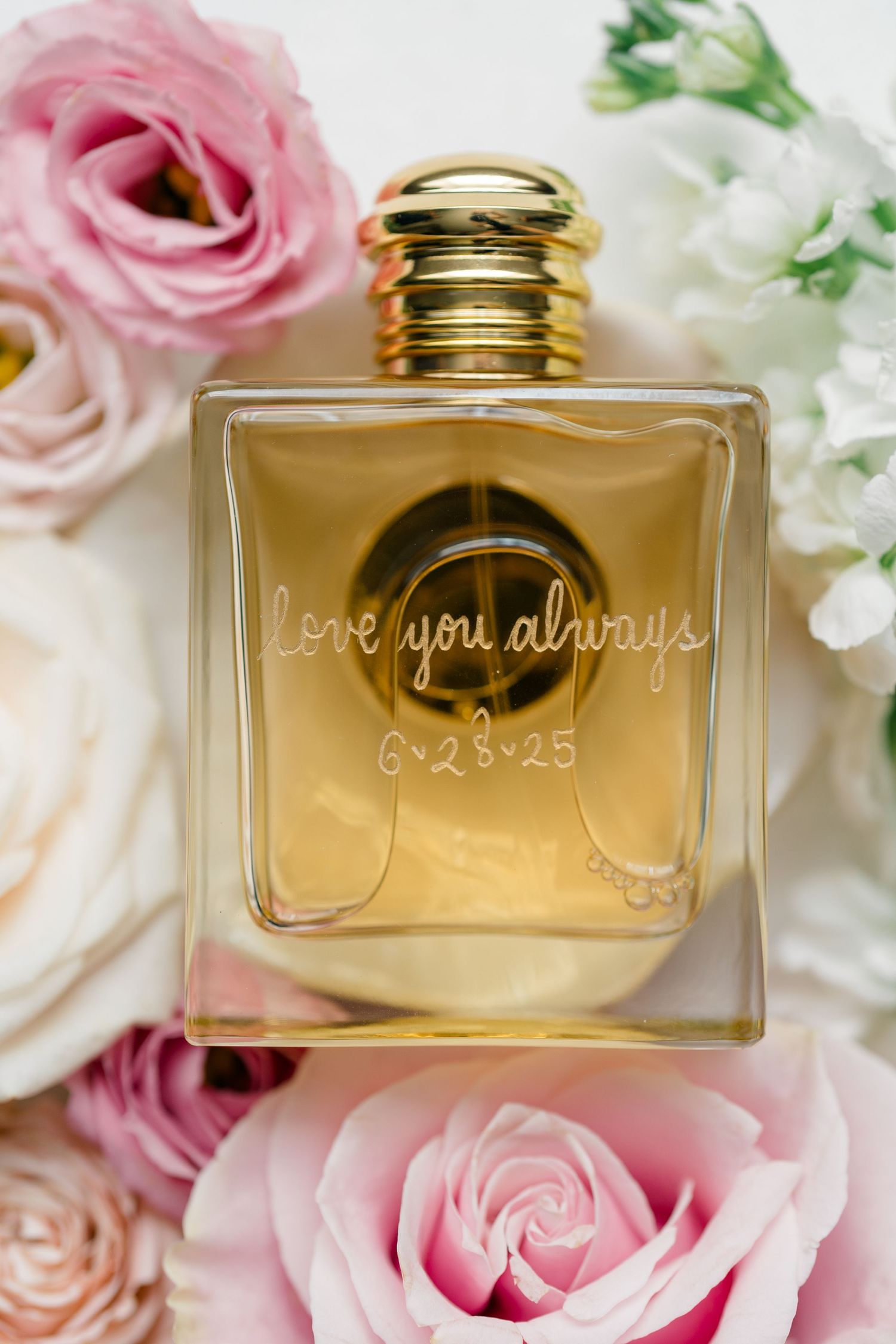 Elegant gold perfume bottle with 'Love you always' engraving nestled among pink roses and white flowers.