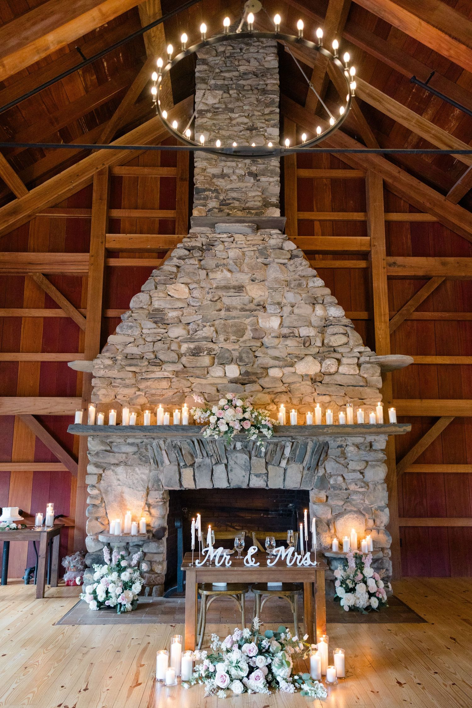 Stone fireplace decorated with candles and white floral arrangements in a rustic barn venue with wood beam ceiling.
