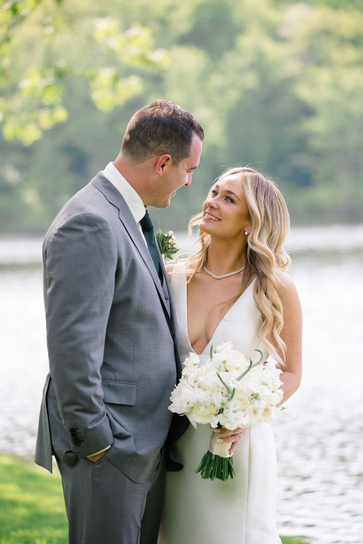 Newlywed couple shares intimate moment by lakeside with bride holding white rose bouquet in elegant wedding attire.