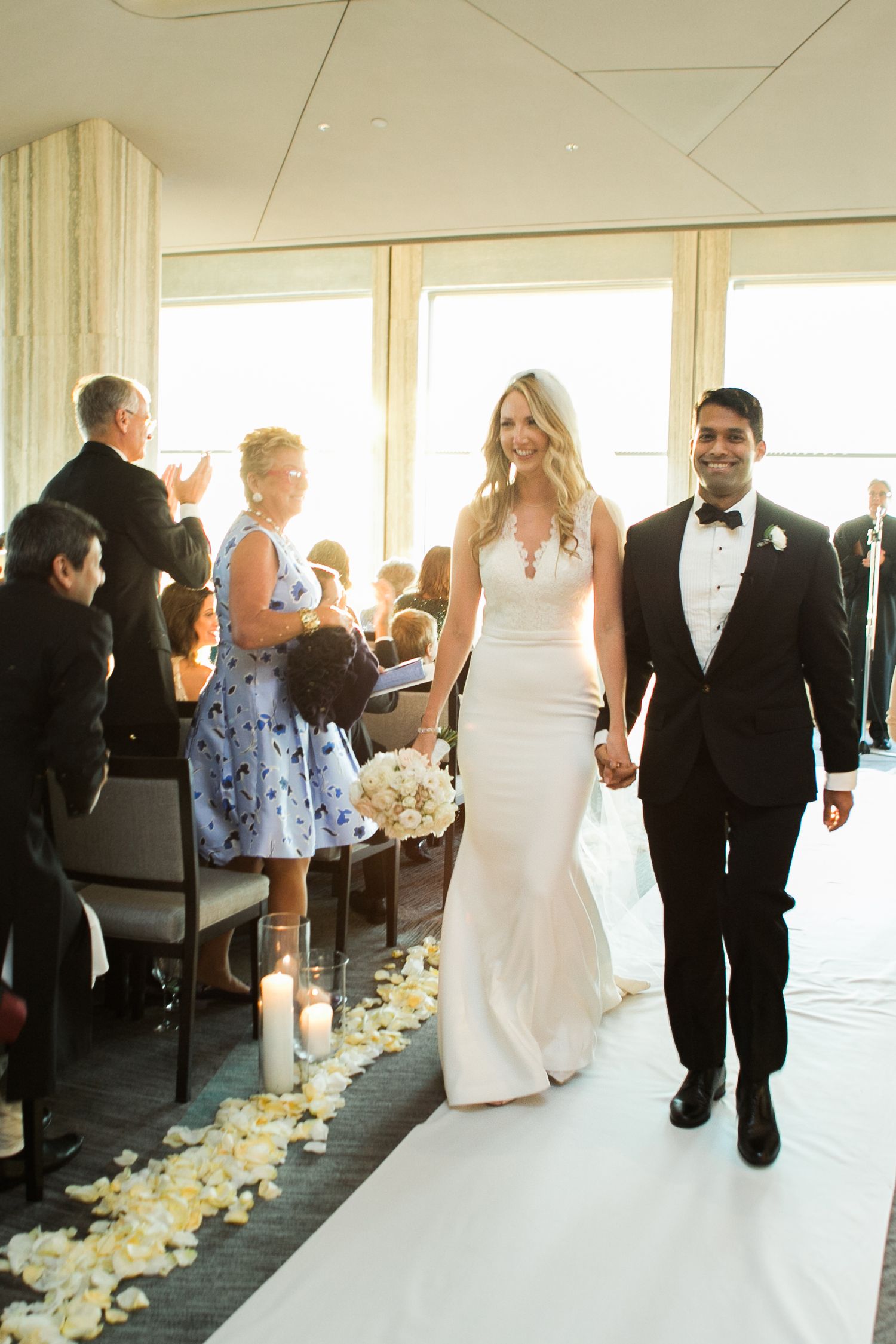 Newlyweds walk down aisle lined with rose petals as wedding guests applaud in sunlit venue.