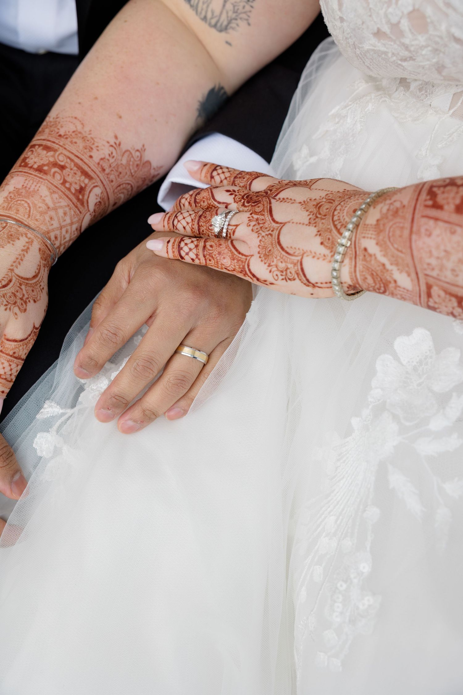 Detailed intricate henna designs and wedding rings displayed on hands against white wedding dress fabric.