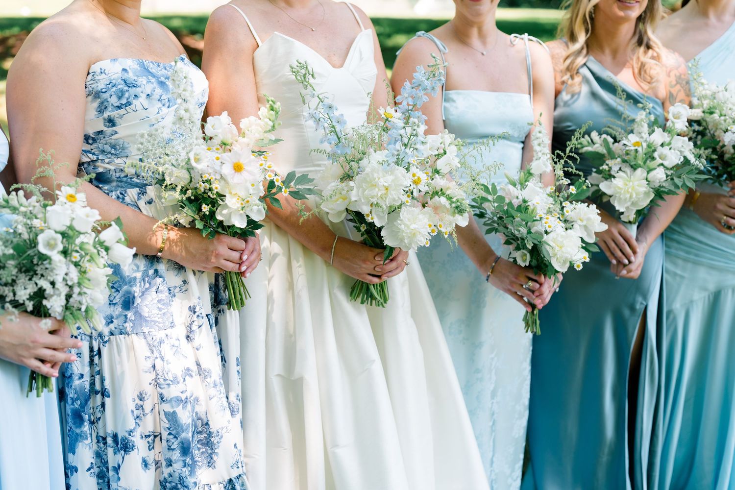 Bride and bridesmaids holding white floral bouquets in blue and white dresses outdoors.