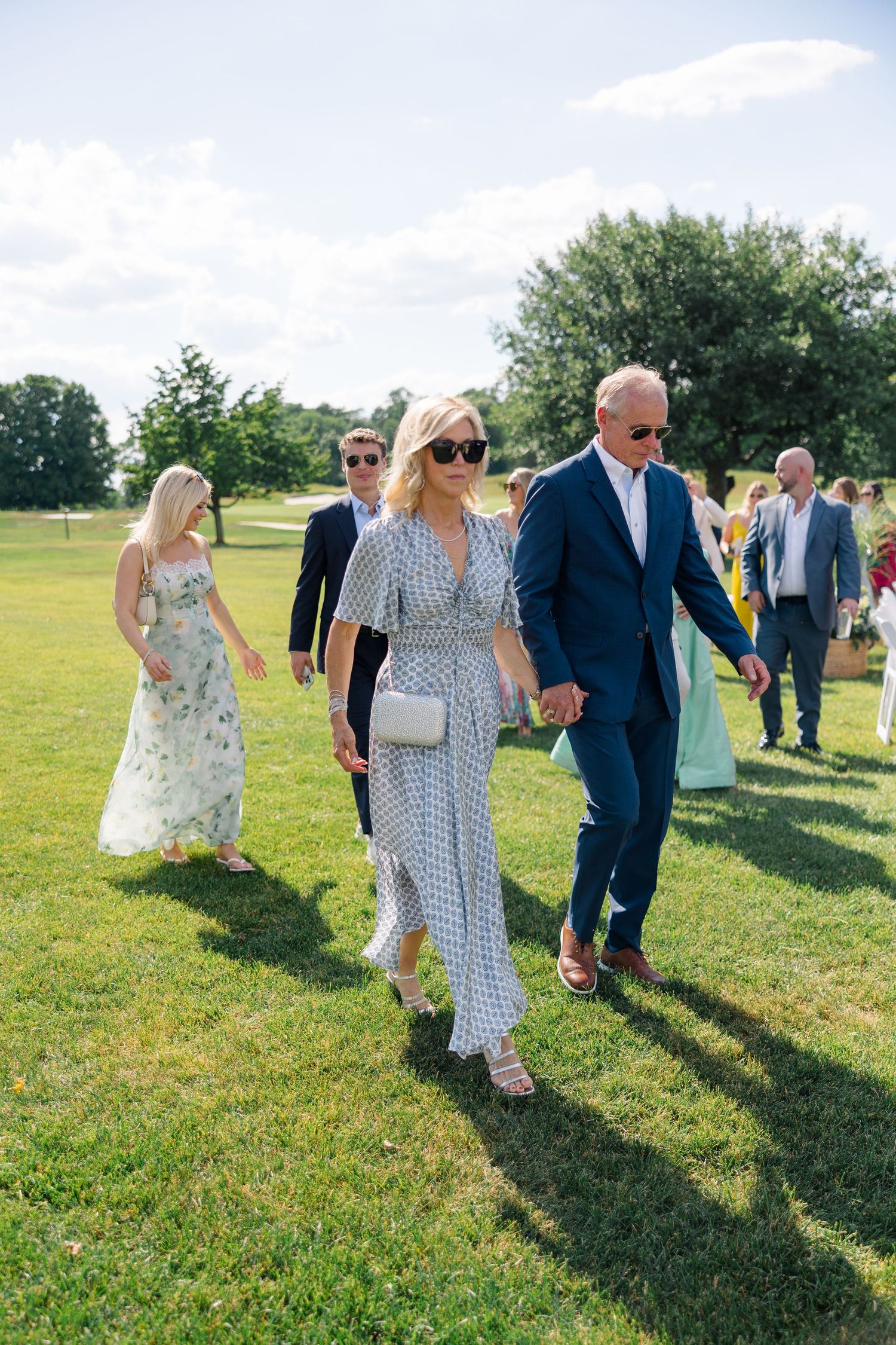 Wedding guests walking across green lawn in summer attire at outdoor garden wedding celebration.