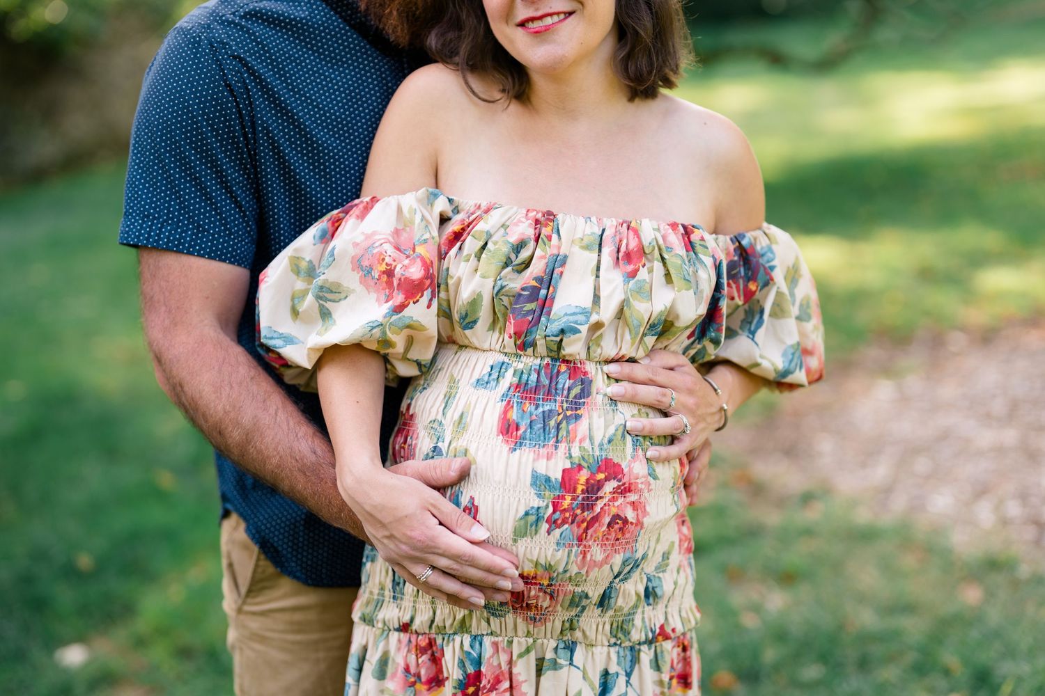Pregnant woman in floral off-shoulder dress cradled by partner outdoors in lush green garden maternity photo.