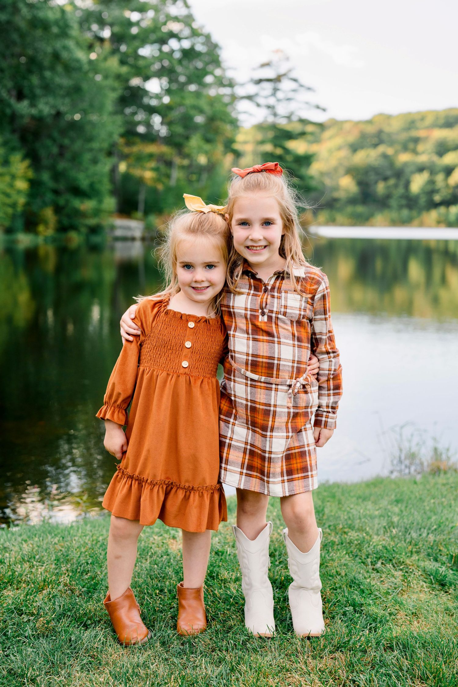 Two young girls in fall outfits pose by a lake, wearing orange and plaid dresses with boots.