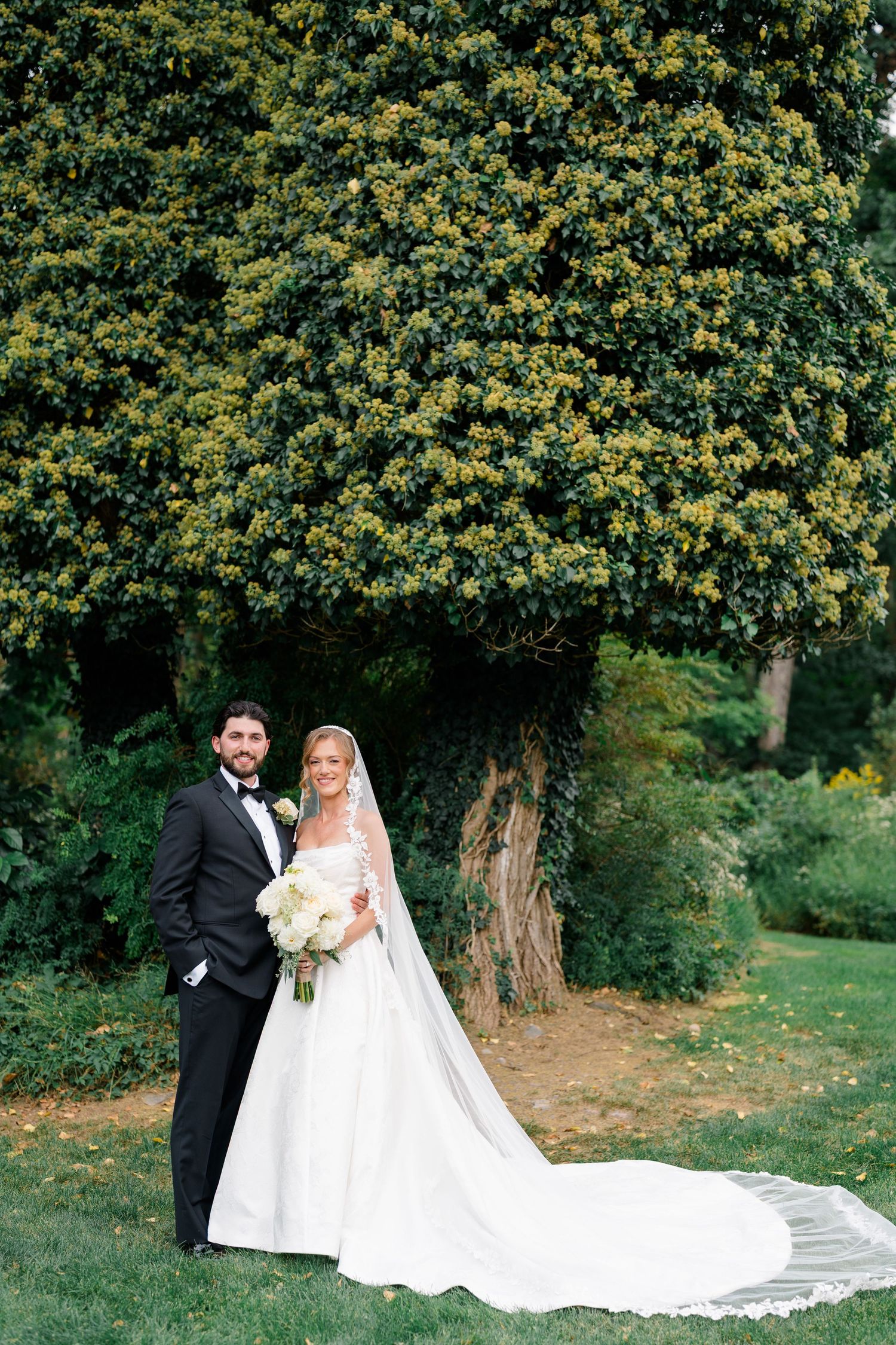 Bride and groom portrait outdoors in lush garden, bride in white ballgown with long veil, groom in black tuxedo.