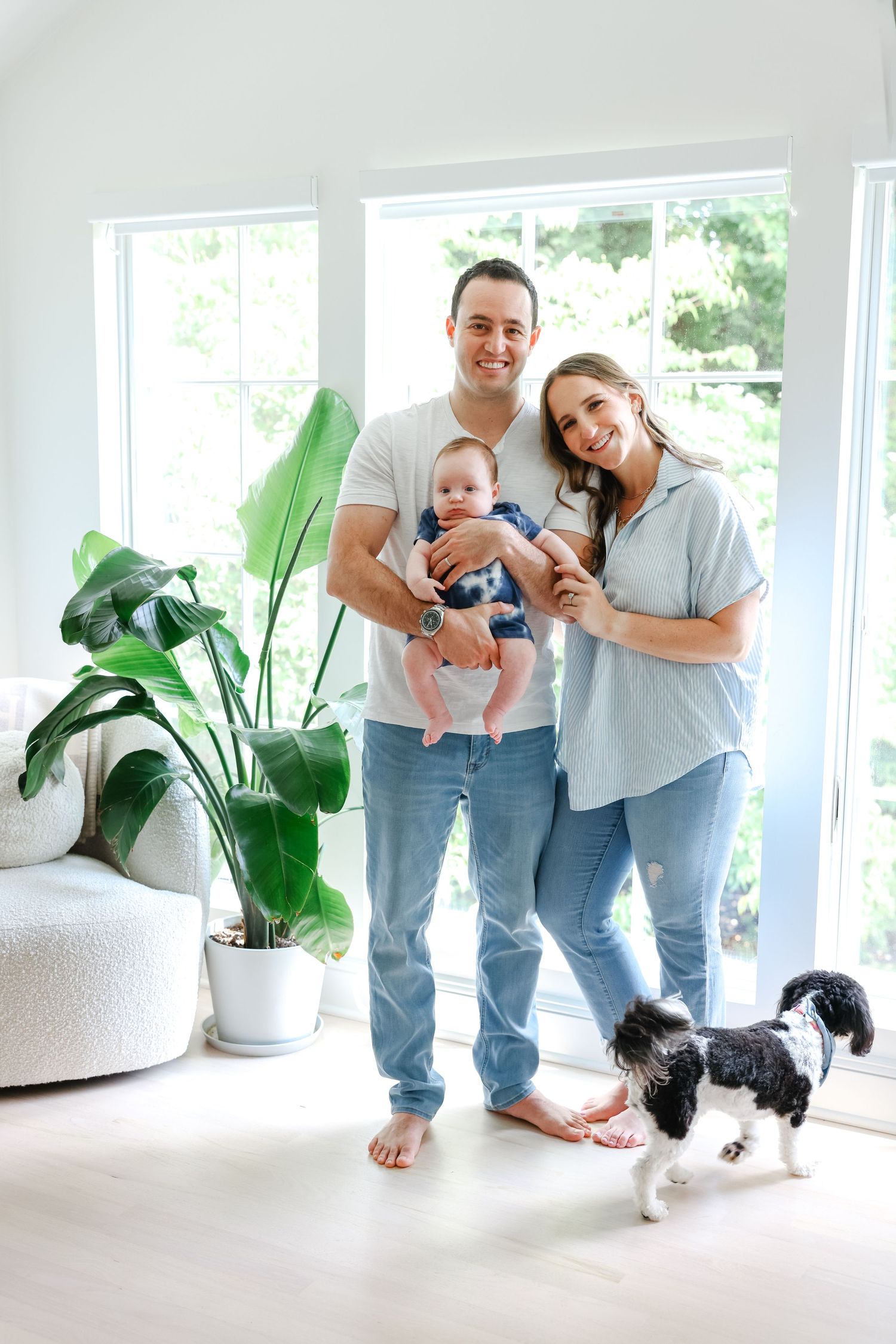 A family stands together in a bright room with large windows, houseplants, and their black and white dog.