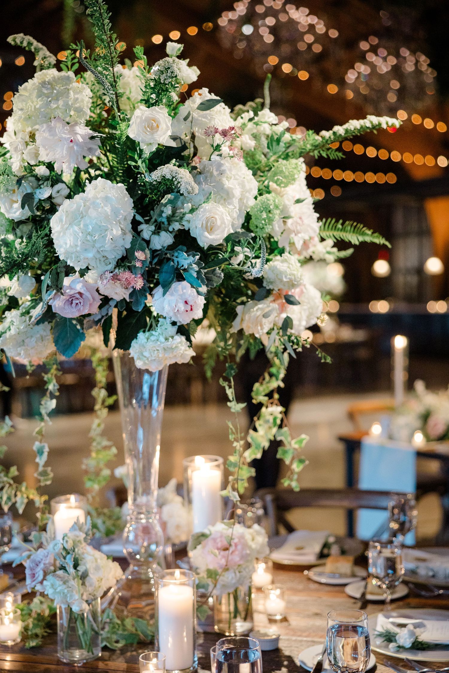 Elegant wedding centerpiece with white hydrangeas and roses in a tall glass vase with glowing candles.