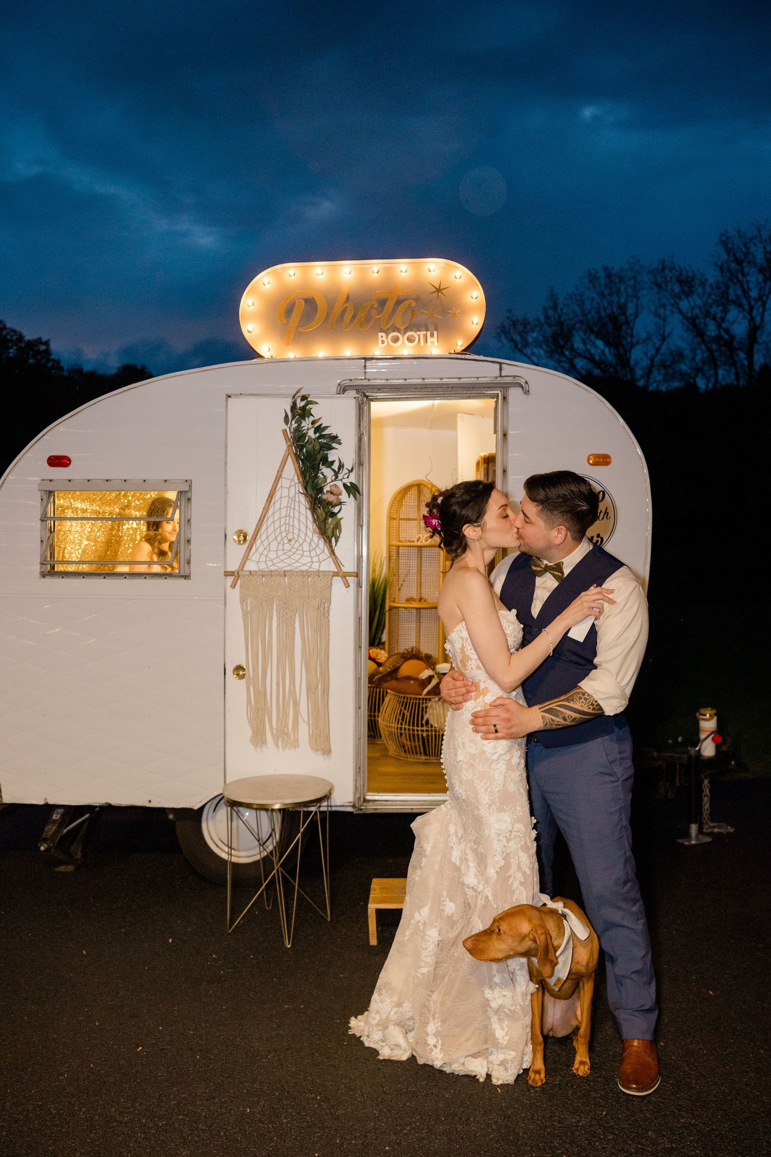 Couple embraces outside illuminated vintage camper with dog at nighttime wedding celebration.