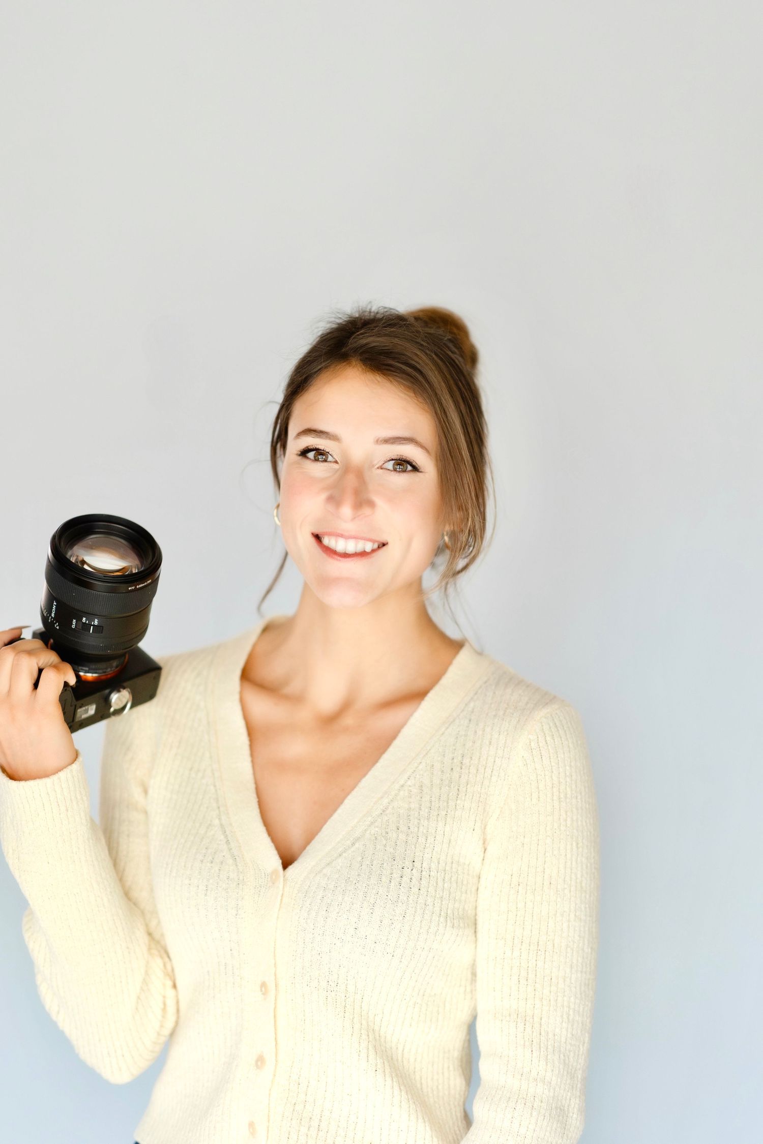 Smiling young woman in cream knit cardigan holding a DSLR camera against a light gray background.