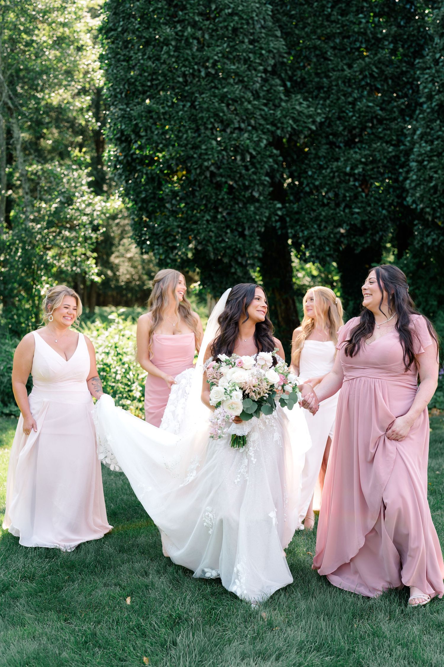 A group in pink and white dresses sits on grass during an outdoor photo session in a garden setting.