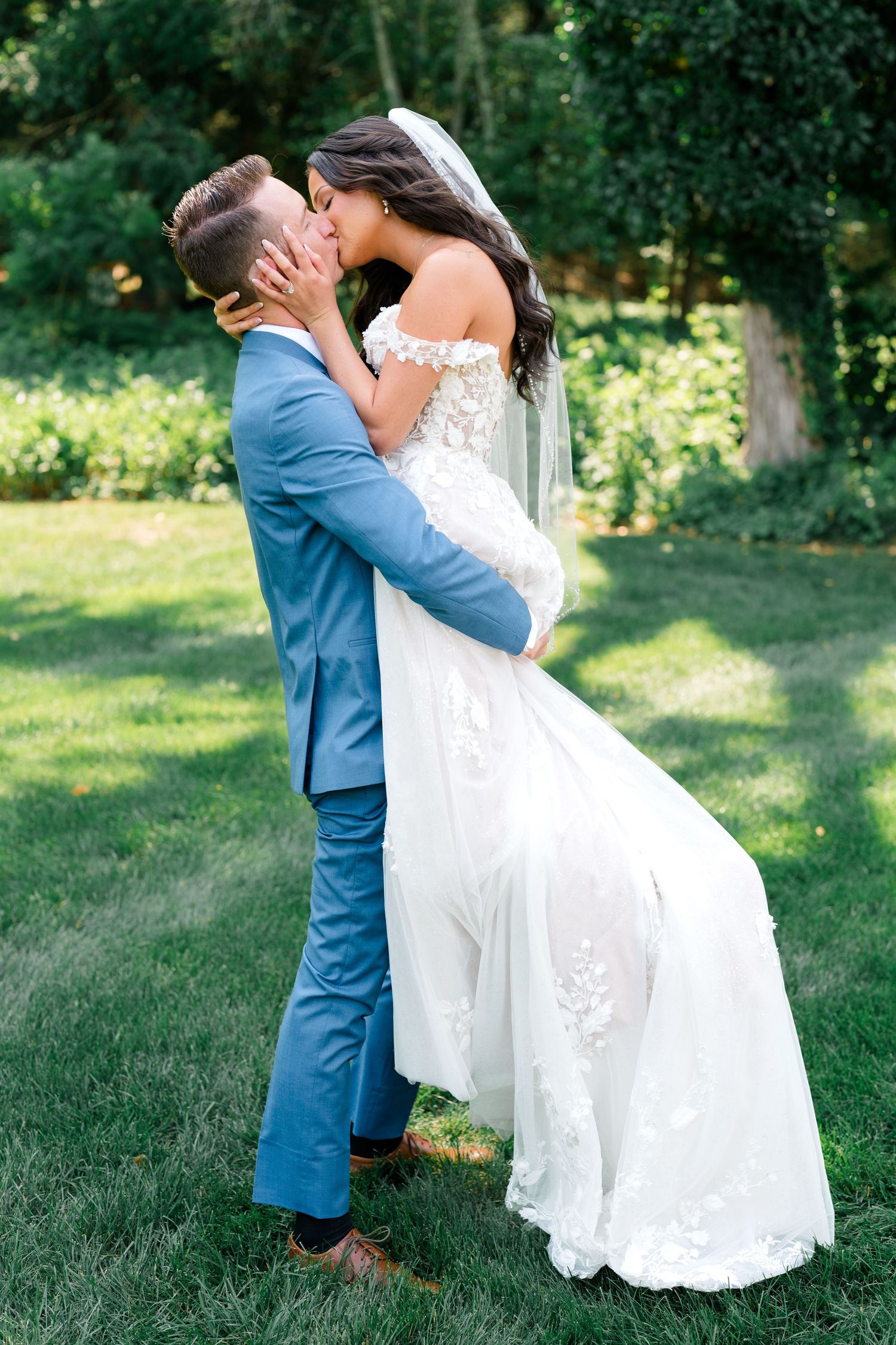 Couple in blue suit and white wedding dress embrace romantically in an outdoor garden setting.