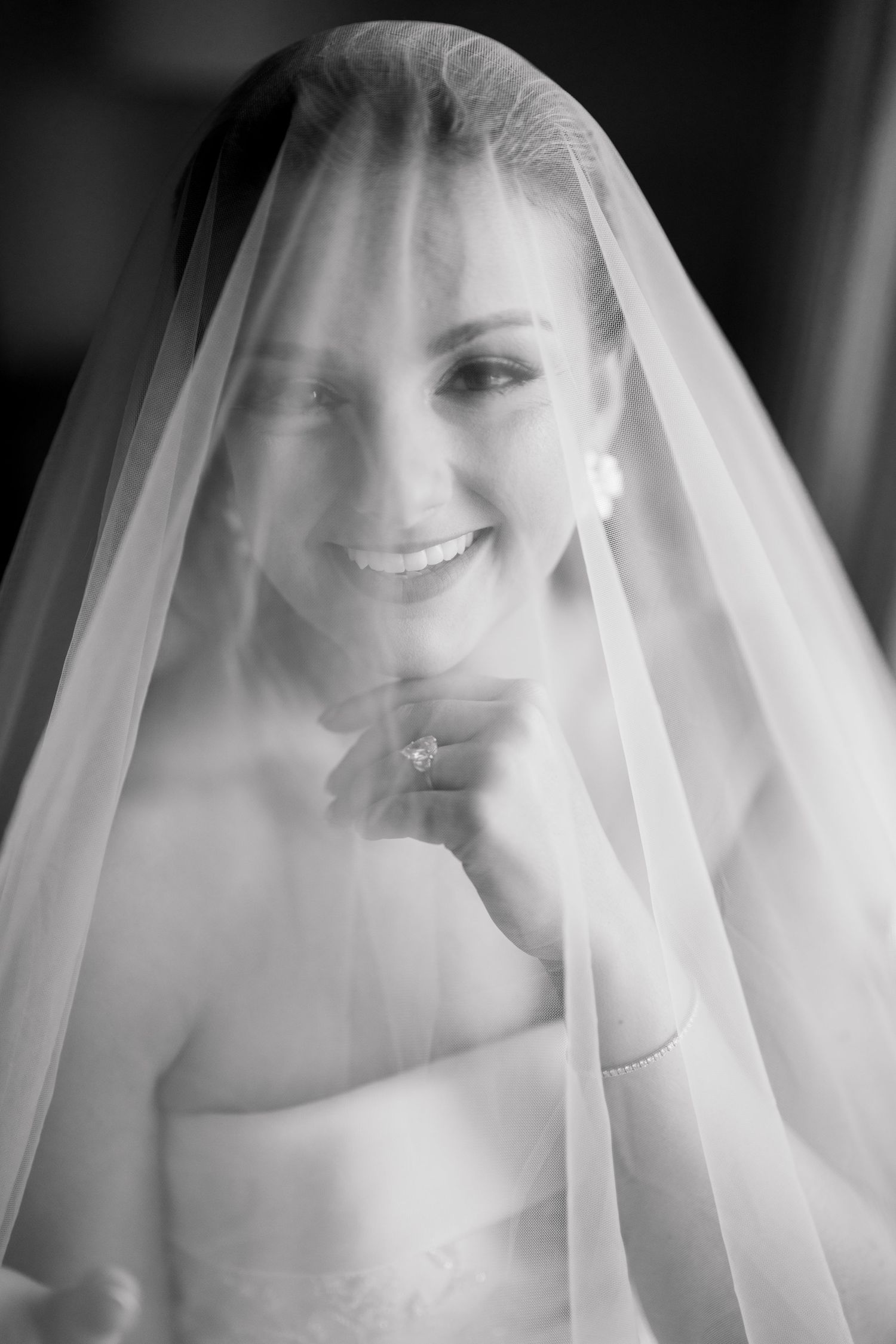 Black and white portrait of a smiling bride wearing a flowing veil and strapless gown, holding her hand to her chin.