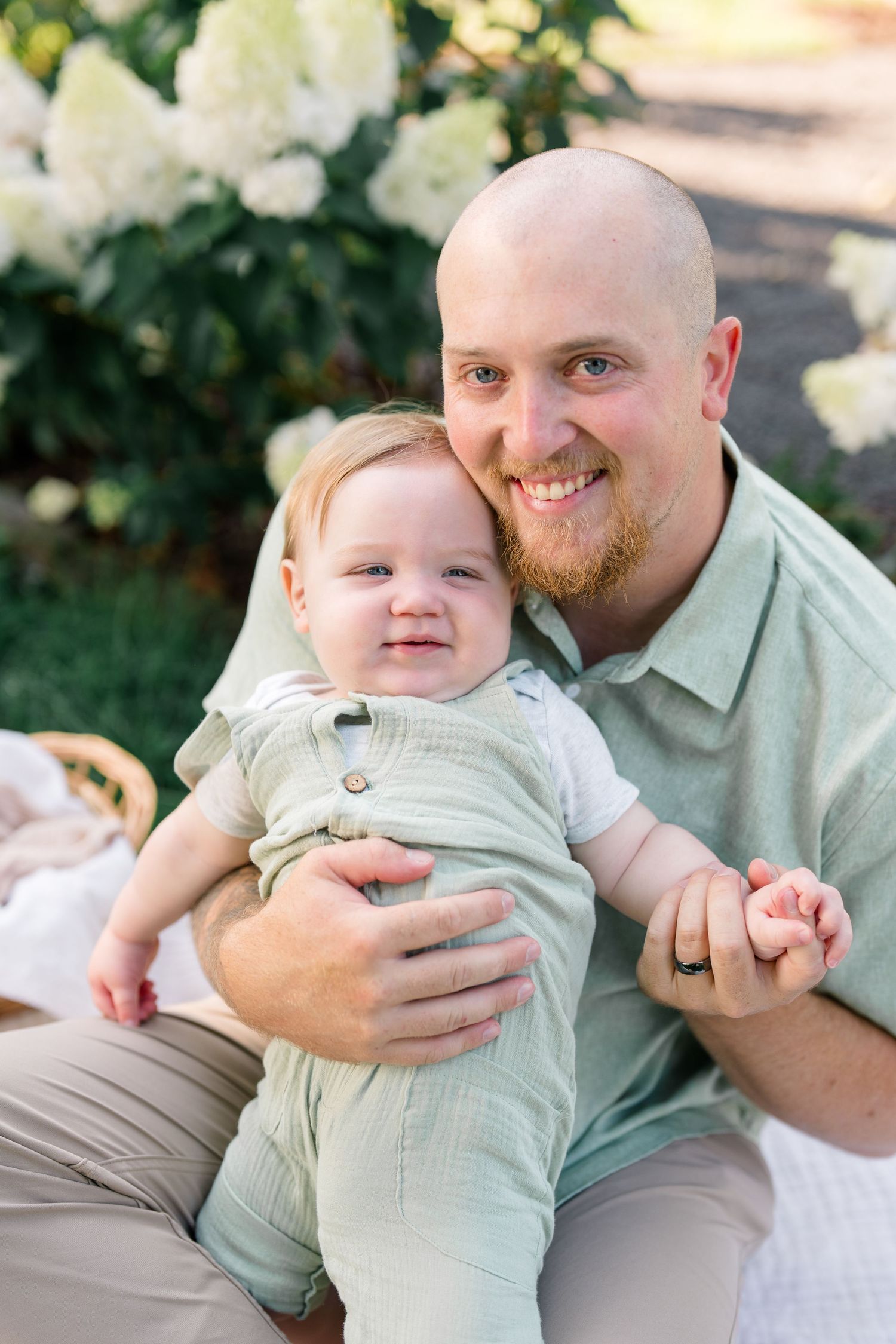 Smiling bald father with beard holds happy baby in green outfit near white flowering bushes.