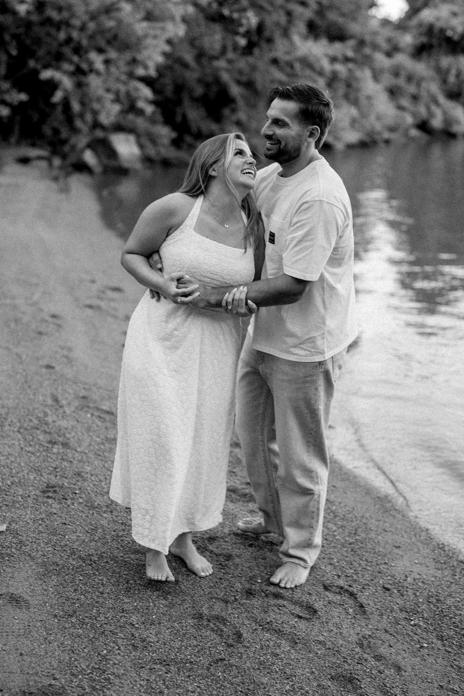 Black and white photo of a couple embracing barefoot by a riverside, woman in white dress laughing.