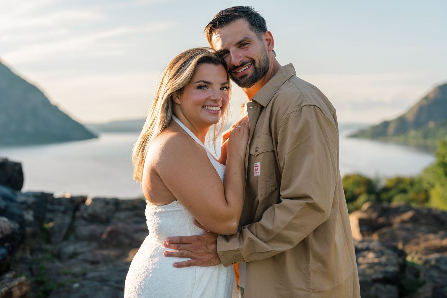 Happy couple embracing outdoors with scenic mountain lake and golden sunset sky in the background.