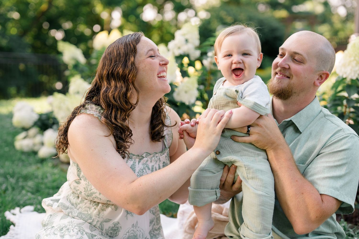 Happy family with bald dad, curly-haired mom, and laughing baby boy in green outfits outdoors near white hydrangeas.