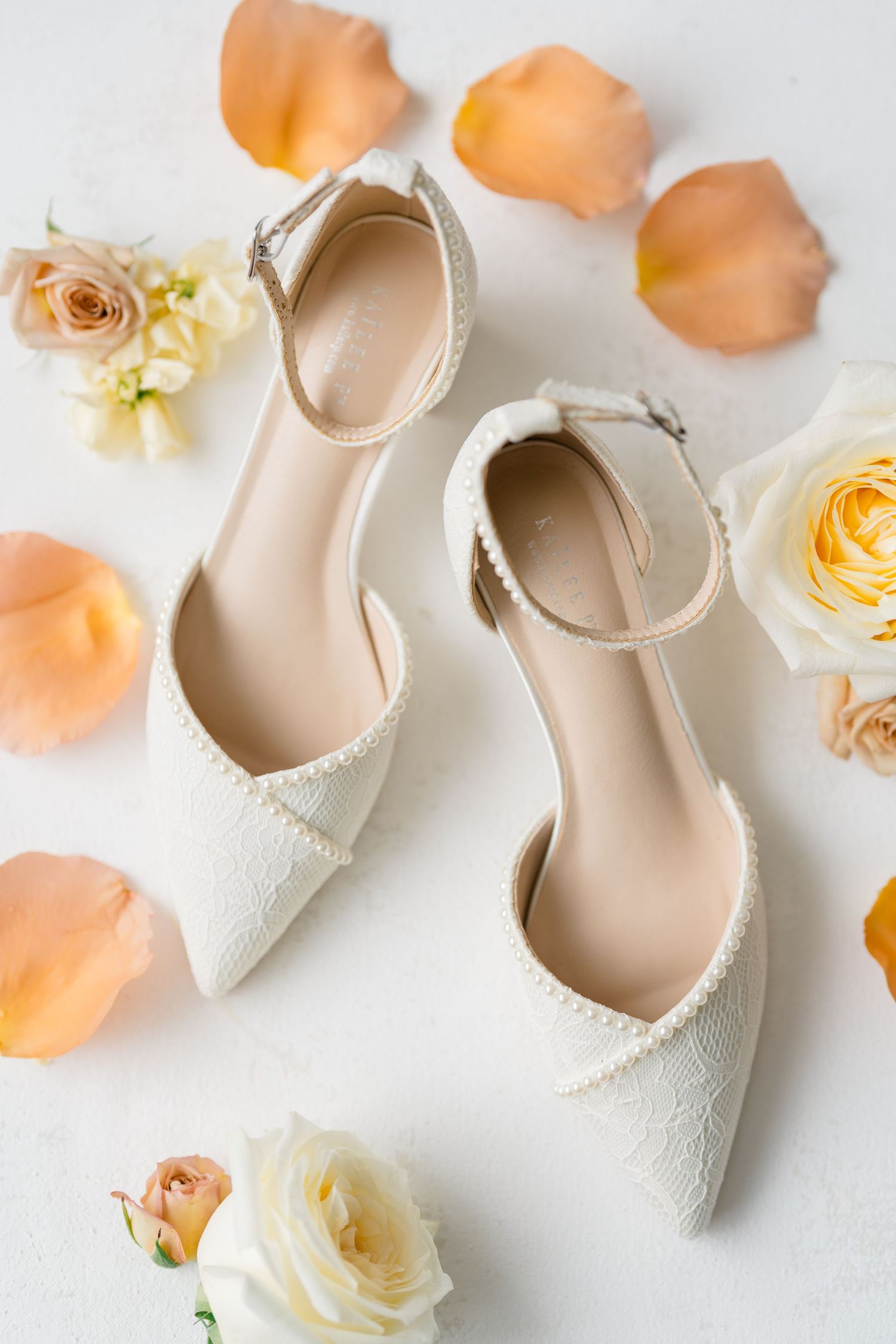 White lace pointed-toe bridal heels with ankle straps surrounded by peach and cream roses and petals.