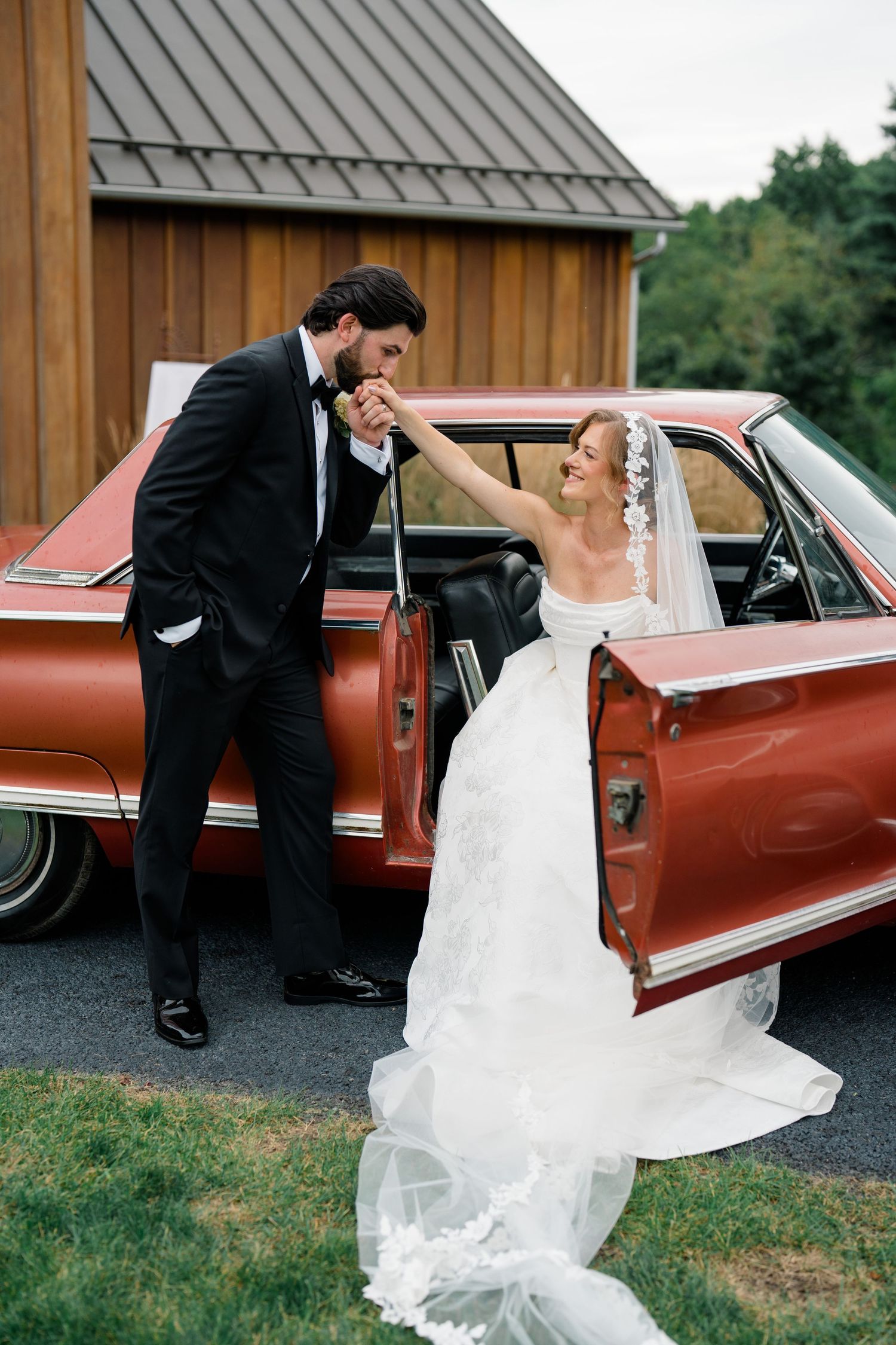 Bride and groom share a romantic moment by a vintage red car, groom kissing bride's hand outside rustic barn.
