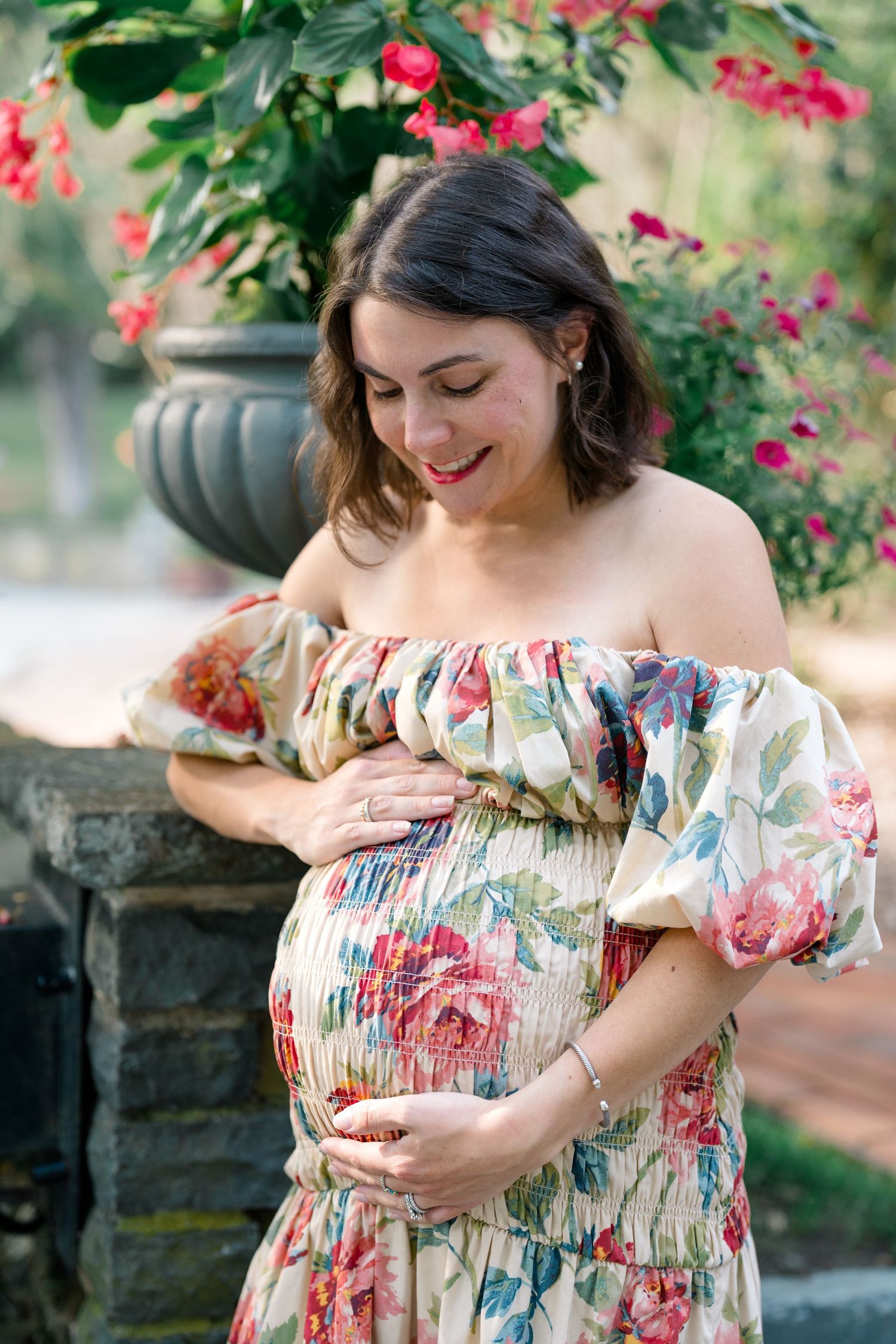 Pregnant woman in floral off-shoulder dress cradling baby bump, smiling outdoors with red flowers in background.
