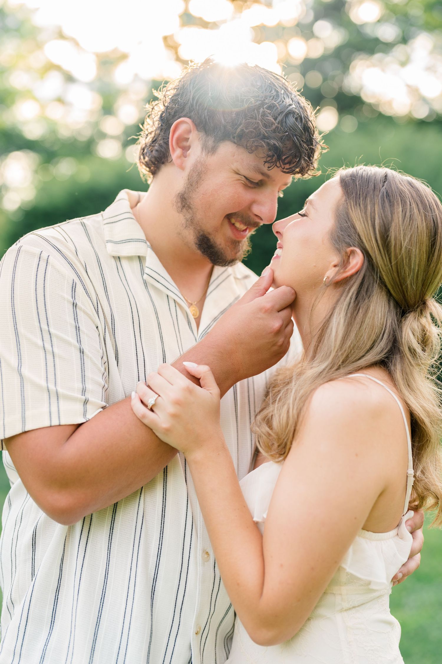 Couple sharing a romantic moment outdoors, man gently holding woman's chin, smiling at each other in golden sunlight.