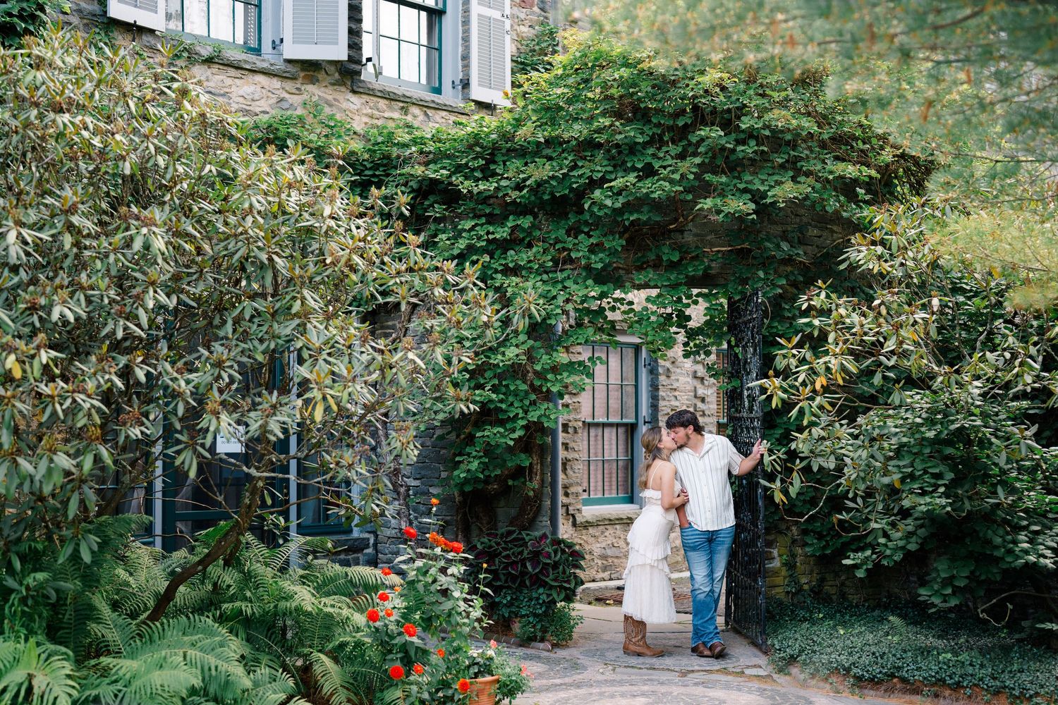 Couple embracing in a lush garden courtyard with ivy-covered stone building and colorful flowers.
