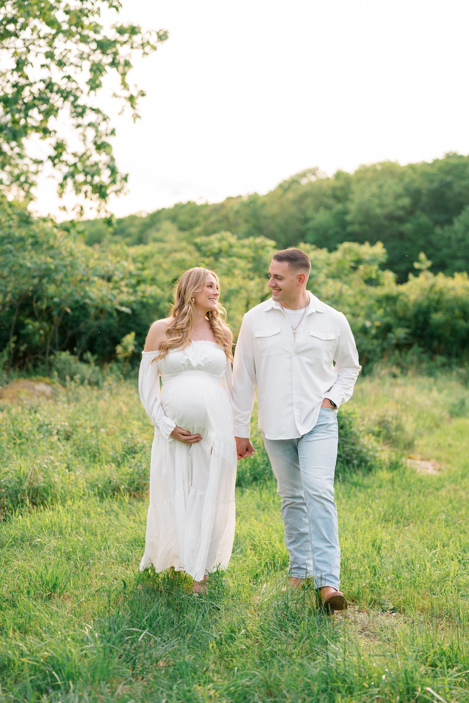 Ethereal maternity photo in flowing white dress against lush green landscape.