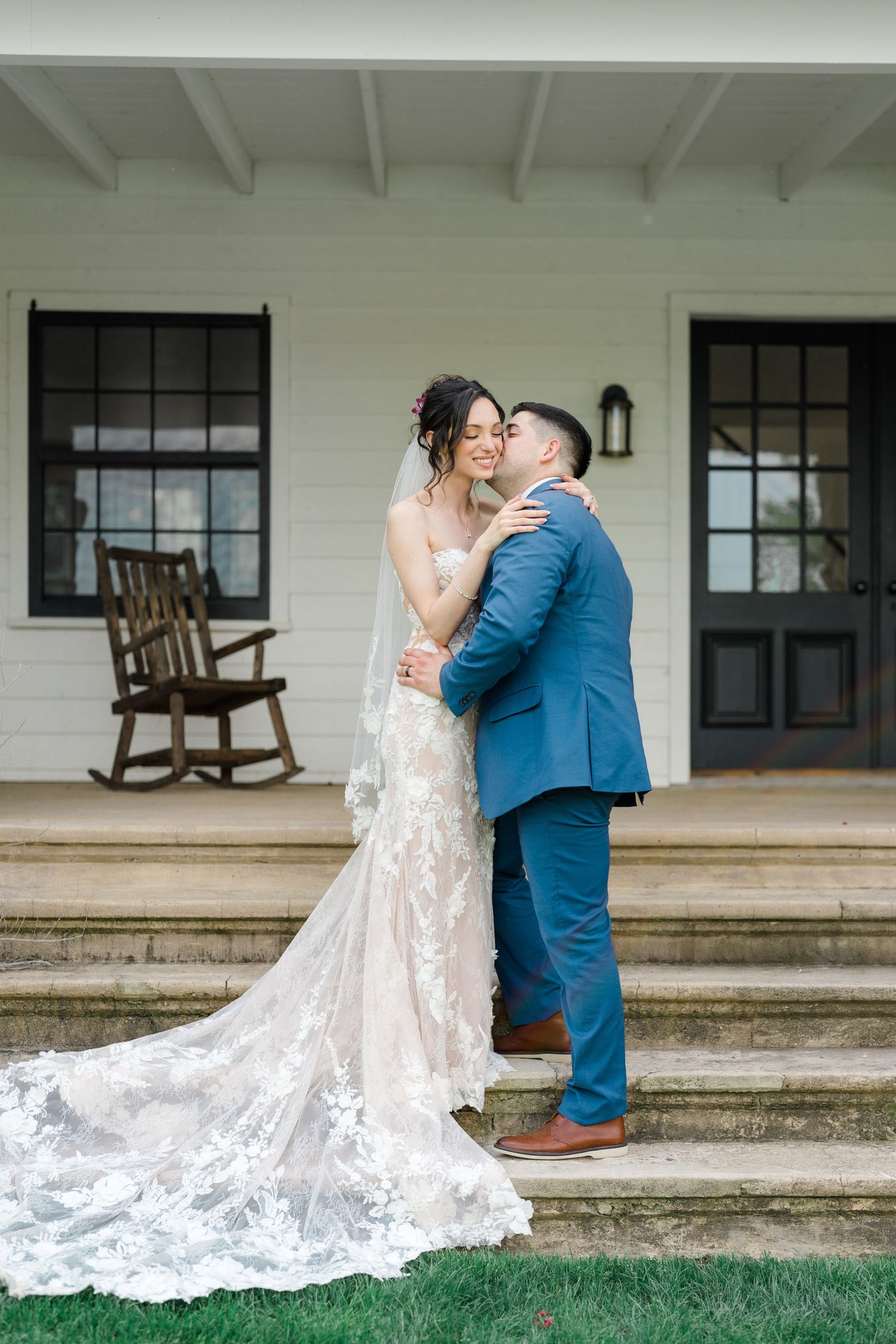 Bride in lace gown and groom in blue suit embrace on white porch steps.
