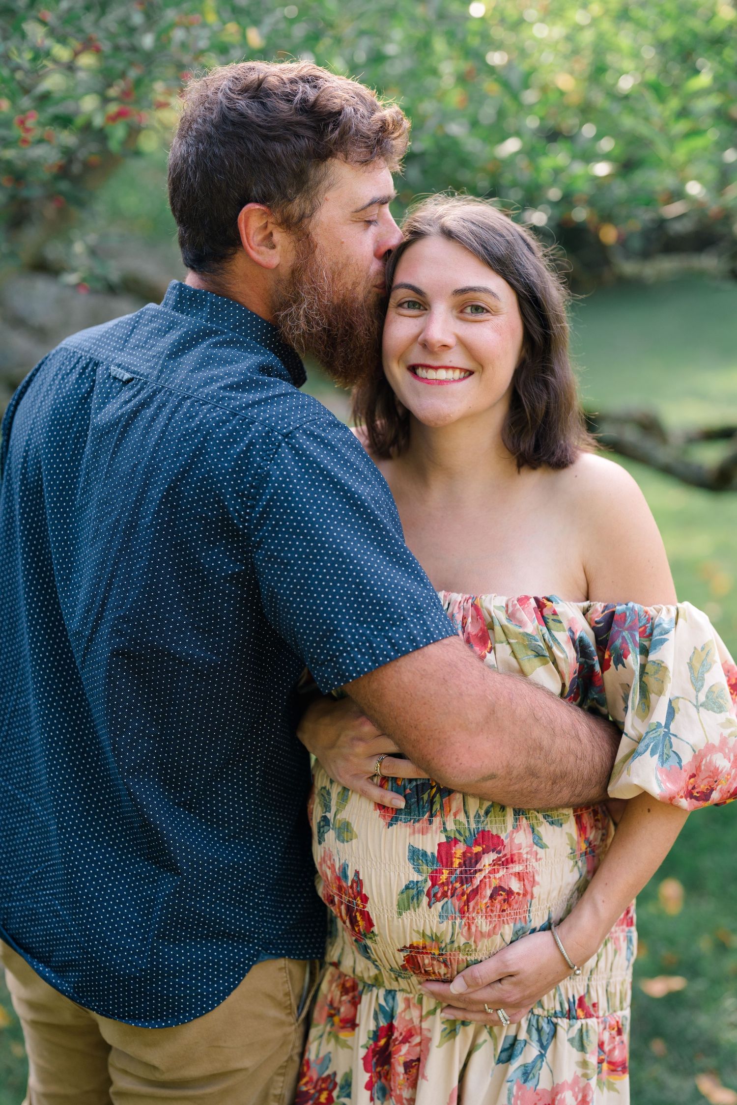A man kisses a smiling pregnant woman in a floral off-shoulder dress outdoors in a lush green garden setting.