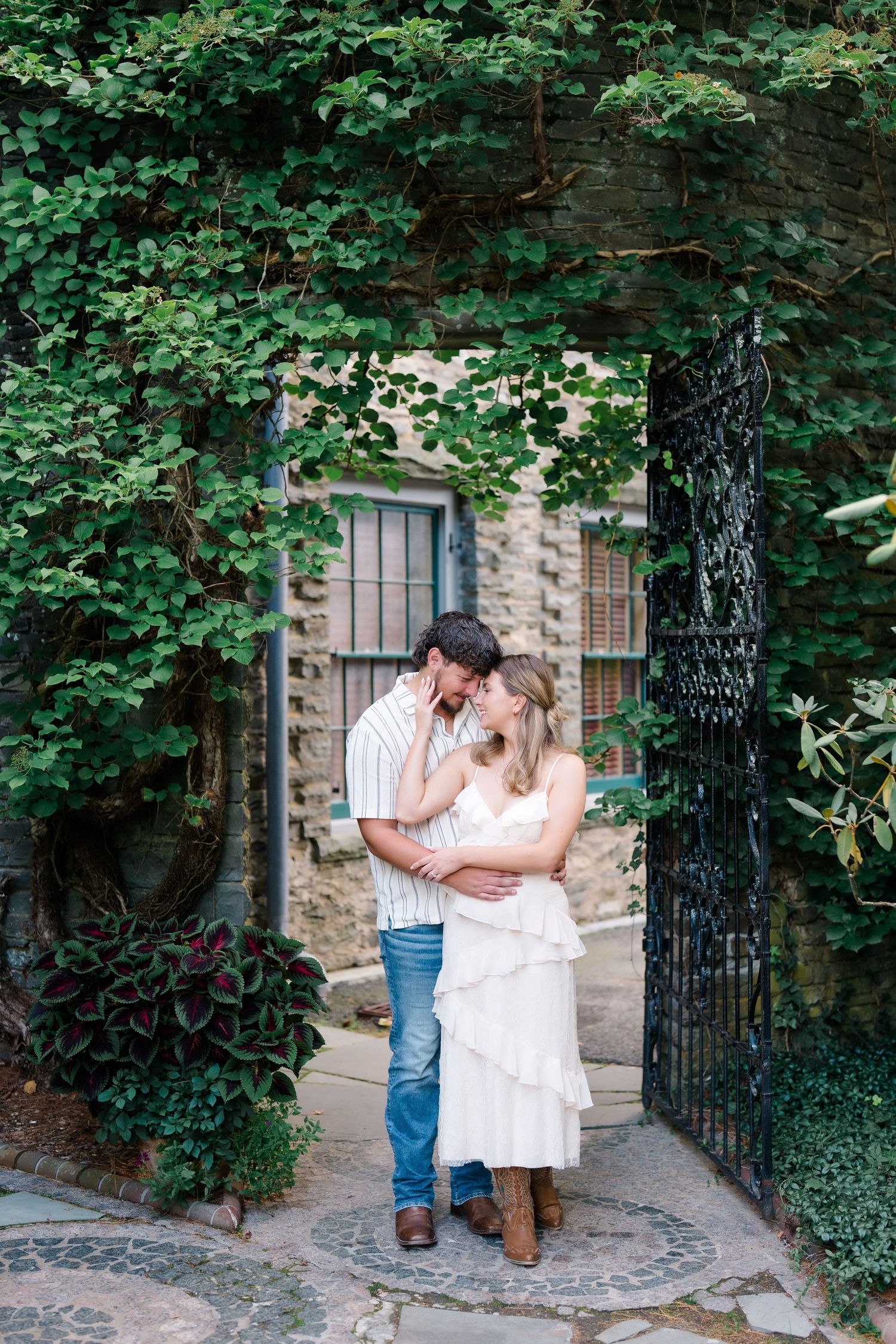 Couple embracing under ivy-covered stone archway with ornate iron gate.