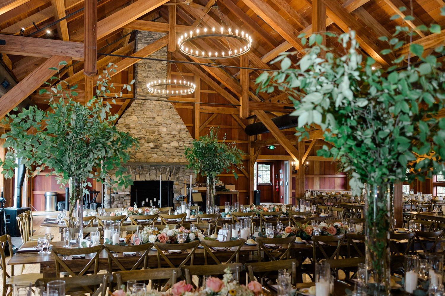Rustic barn wedding reception with tall greenery centerpieces, long tables, chandelier lighting, and stone fireplace backdrop.