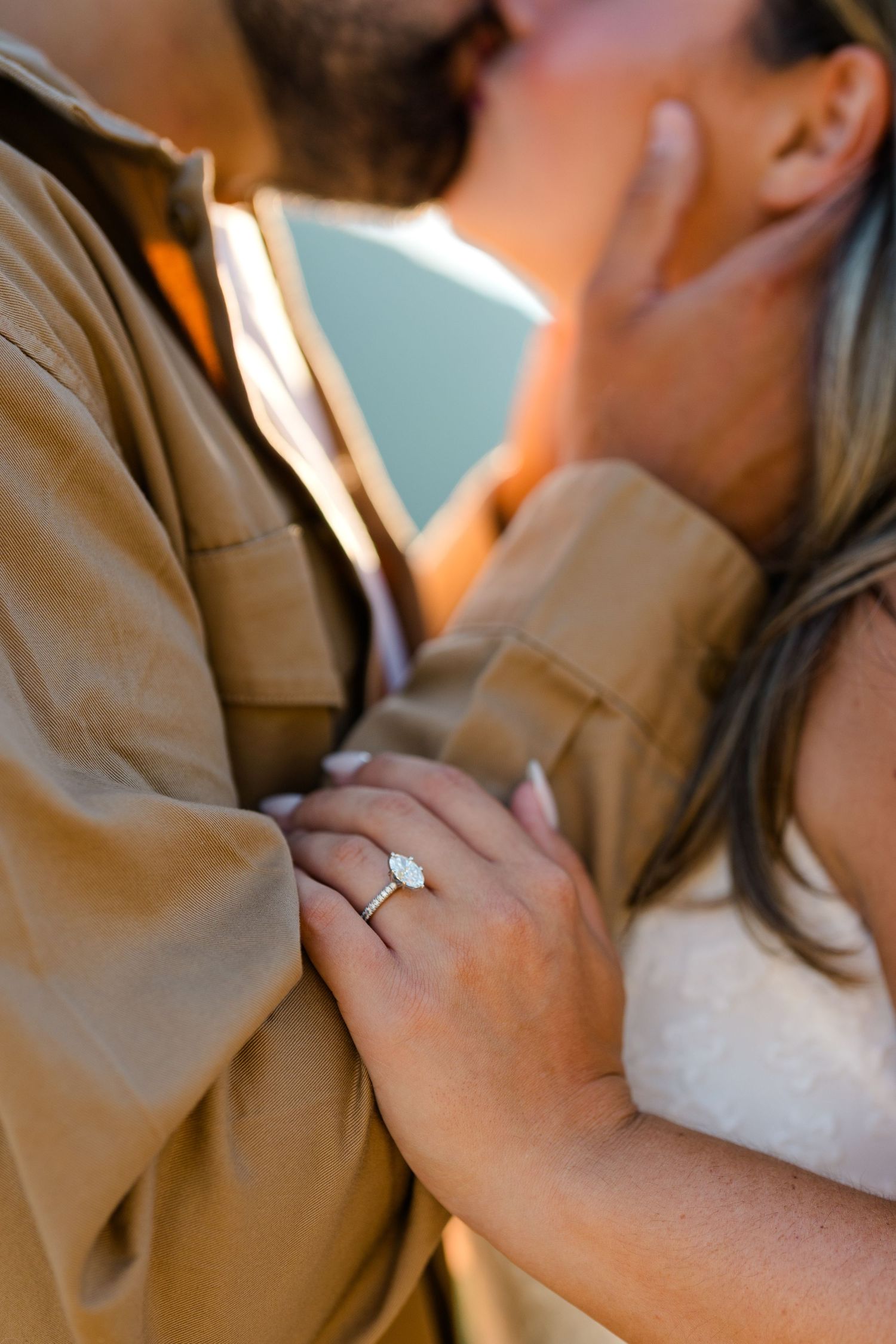Couple embracing with woman showcasing elegant diamond engagement ring on man's arm in warm golden light.