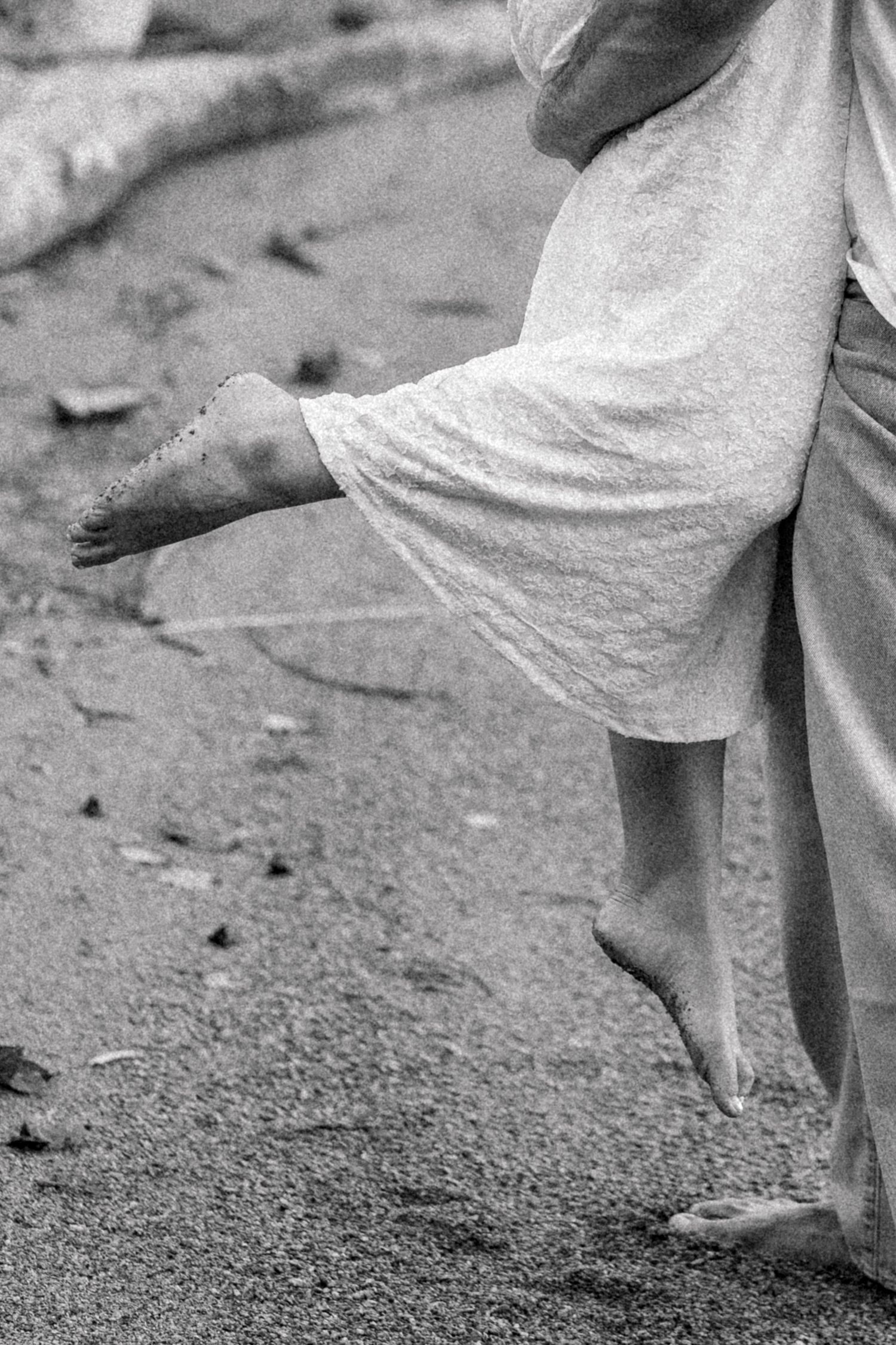 Black and white photo of bare feet walking on sandy beach, wearing a flowing white dress, captured mid-stride.