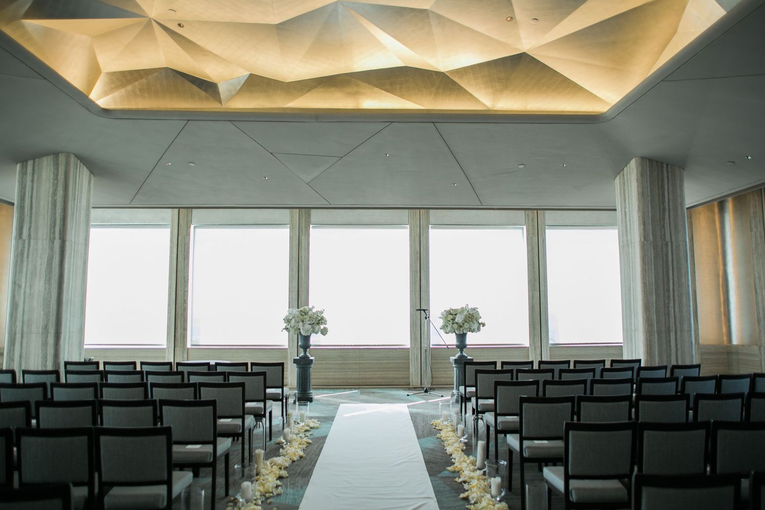 Modern wedding ceremony venue with geometric ceiling, rows of black chairs, and floor-to-ceiling windows overlooking natural light.