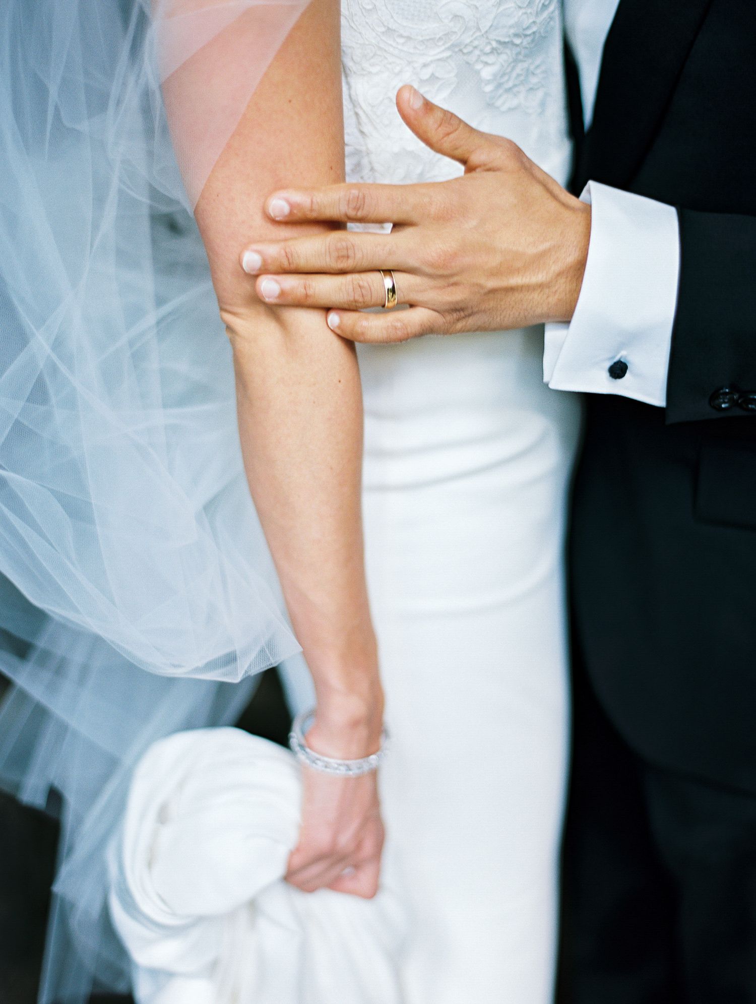 Close-up of hands and wedding ring detail against white dress and black tuxedo.