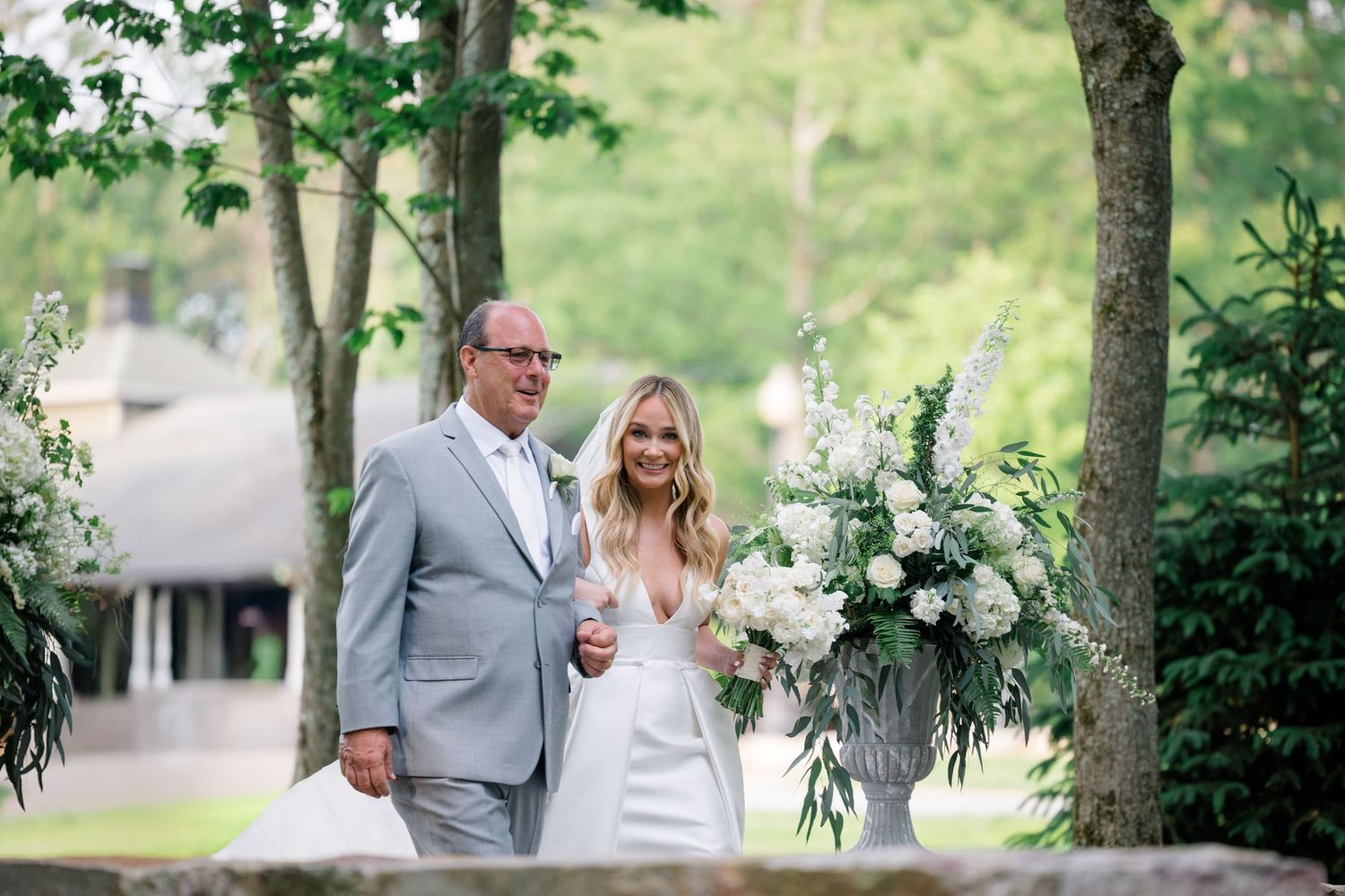 A couple walks together on a wooded path, the bride holding a lush white floral bouquet with greenery.